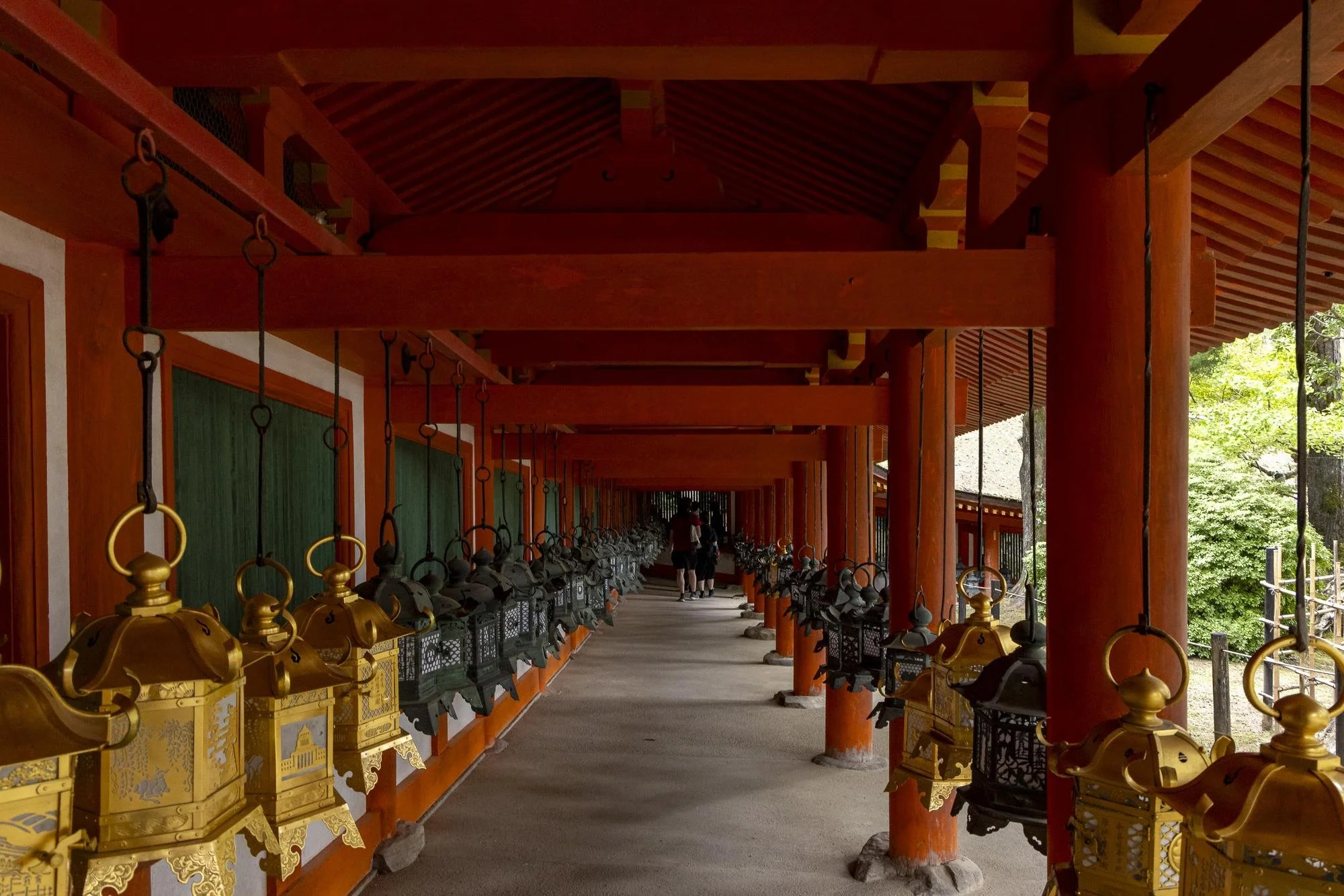 A covered walkway with wooden beams and posts, lined with traditional Japanese lanterns on both sides, leading to a couple walking in the distance, surrounded by greenery.