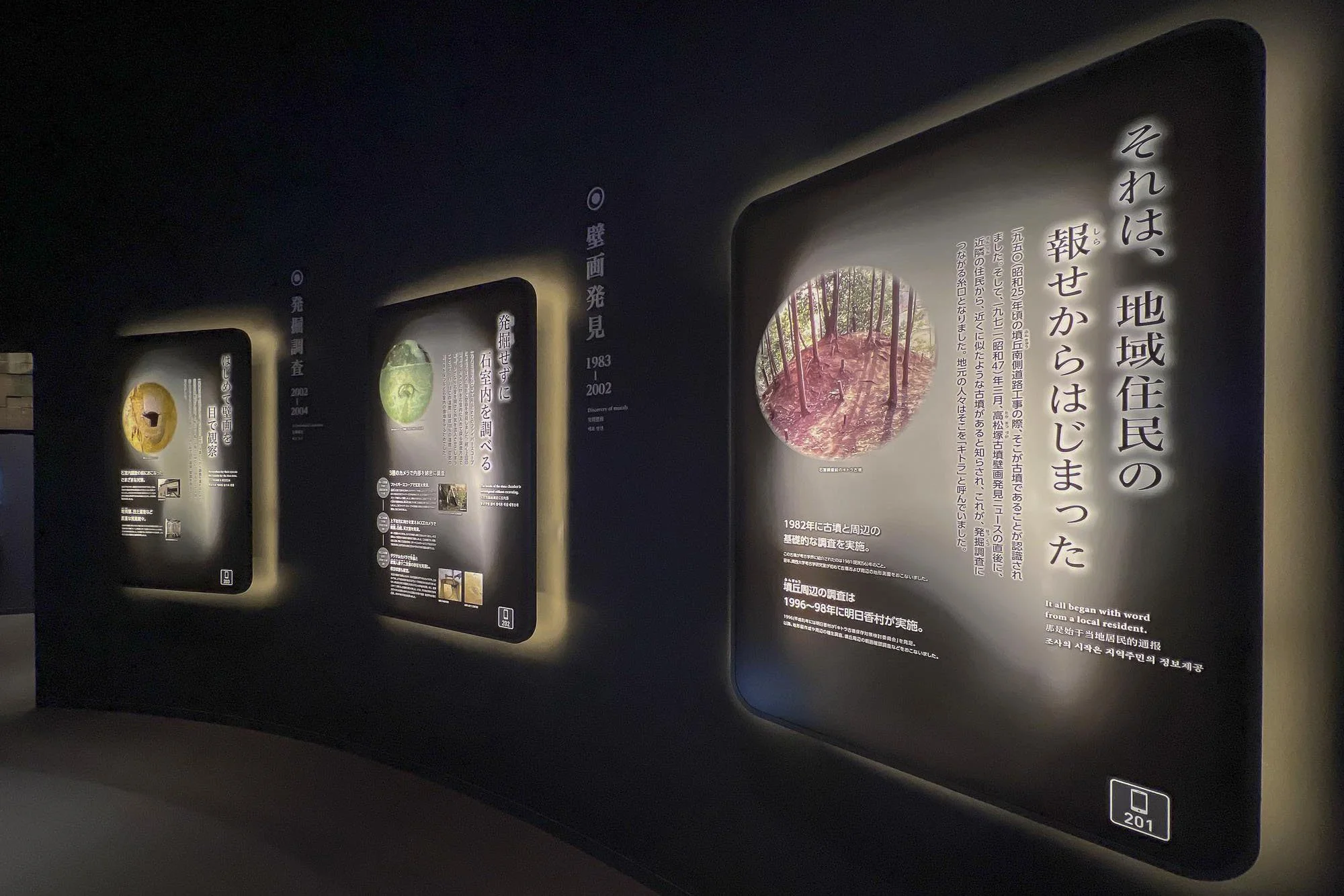 Exhibit panels with Japanese and English text and images about regional history and archaeology, illuminated on a dark wall.