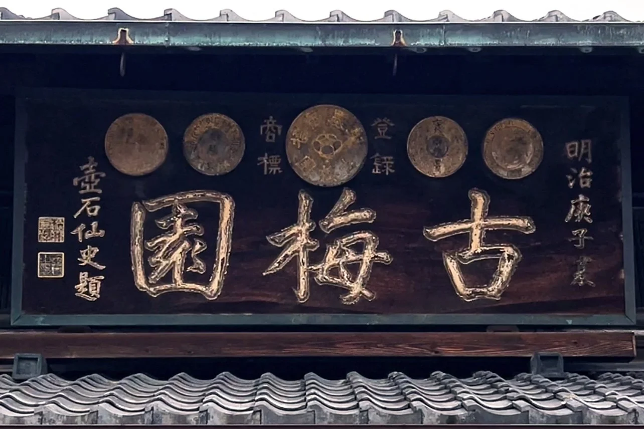 Wooden sign with large Chinese characters and gold coins displayed above, mounted on a traditional tiled roof.
