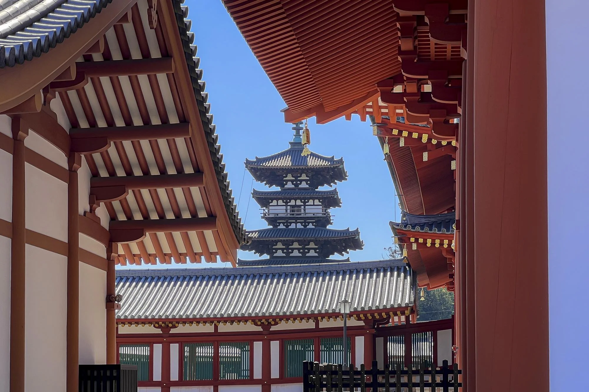 View of traditional Japanese temple architecture with a multi-tiered pagoda in the background, red and white wooden structures, and a clear blue sky.