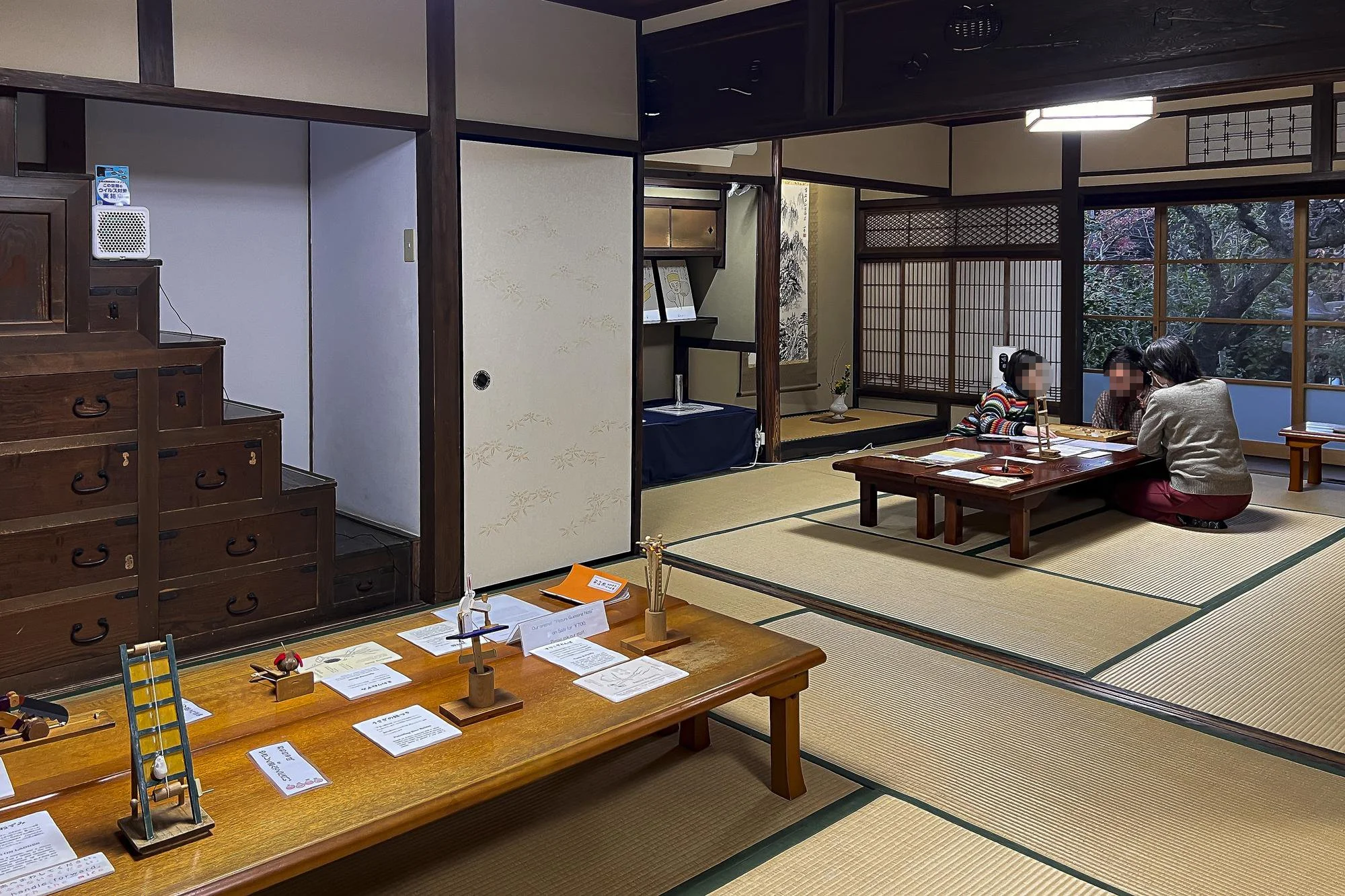 Interior of a traditional Japanese room with tatami mats, a low wooden table, and sliding shoji doors. Three people are sitting on the floor, having a conversation. Decor includes a hanging scroll and a small flower arrangement, with an outdoor garde