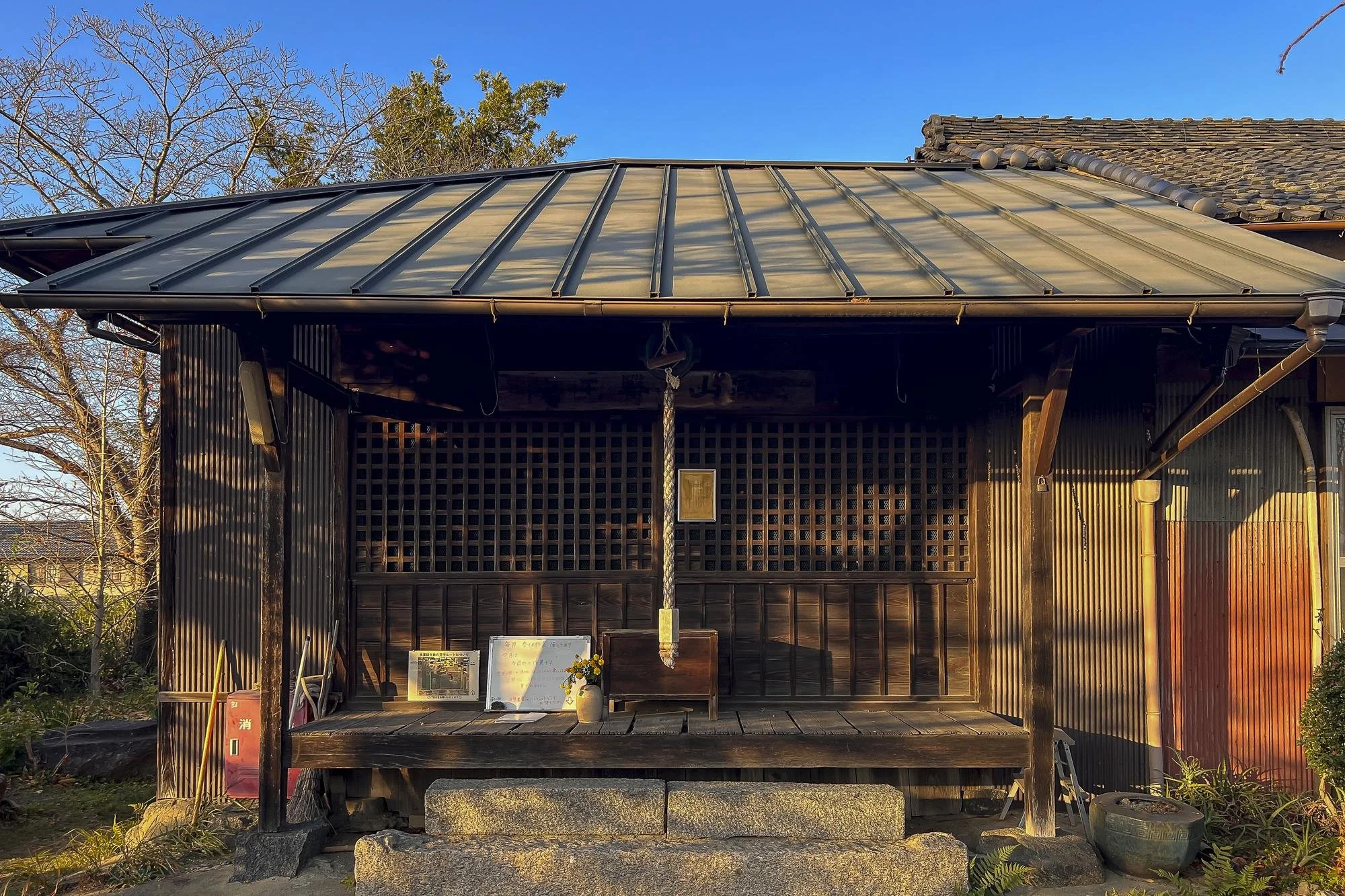 Traditional Japanese wooden shrine with a raised platform, a bell hanging in the center, and a small offering area with a vase of flowers and plaques. The shrine is outdoors on a stone foundation with a metal roof, surrounded by trees and under a cle