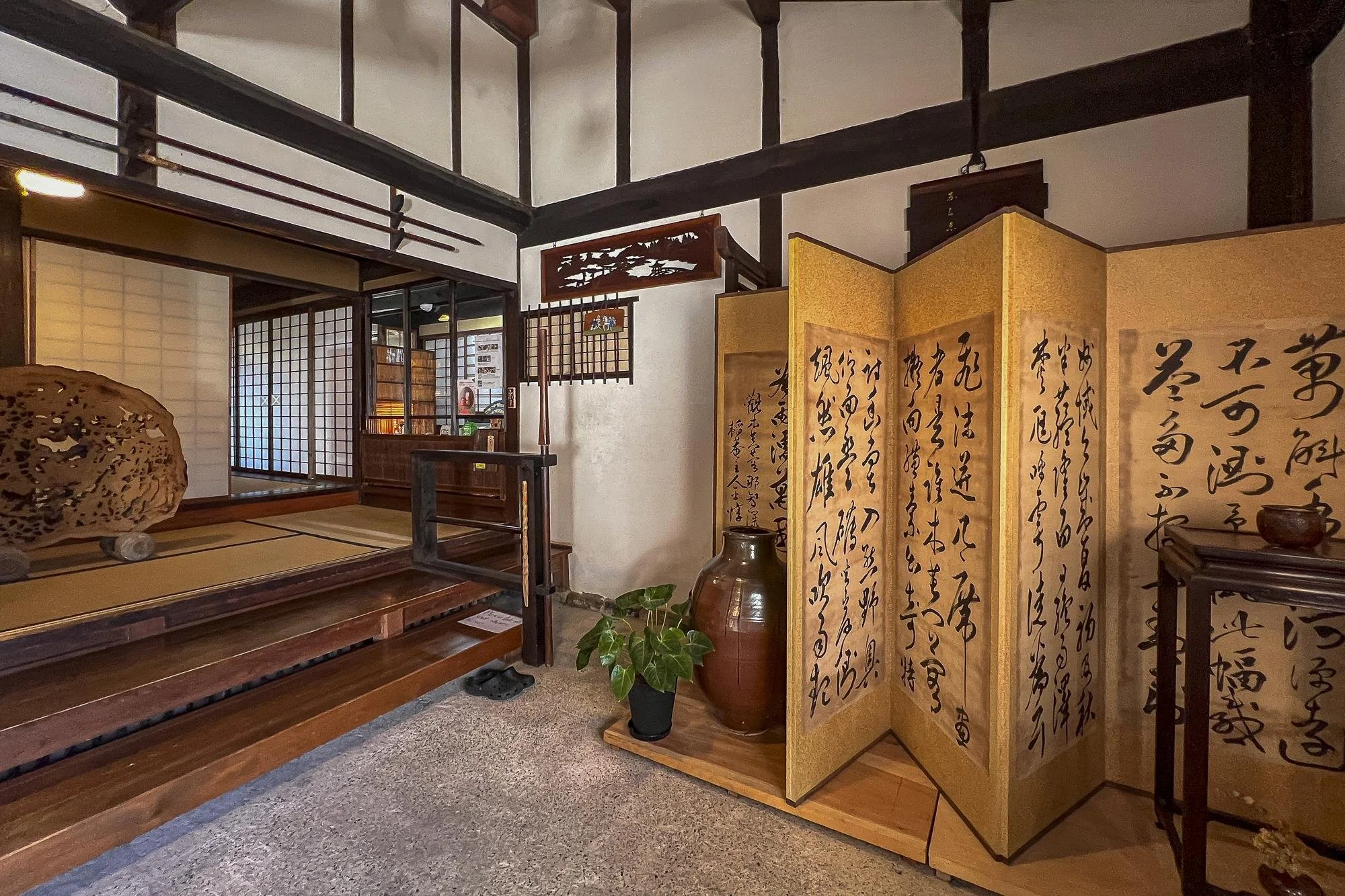 Interior of a traditional Japanese room with wooden accents, shoji sliding doors, a folding screen with calligraphy, a large pottery vase, and a potted plant.