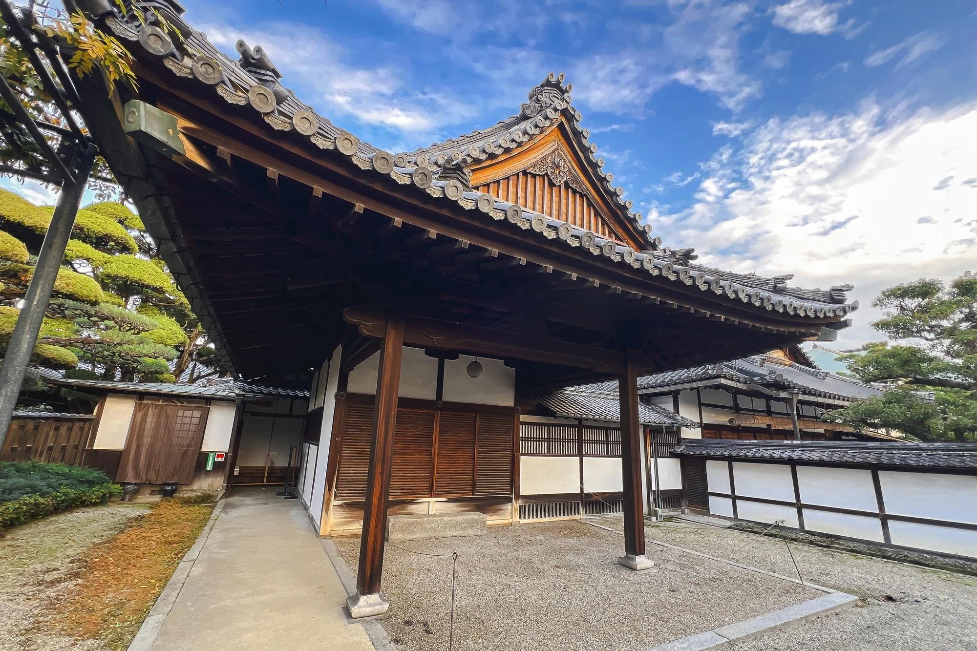 Traditional Japanese wooden building with curved roof, surrounded by trees and a gravel courtyard, under a blue sky with scattered clouds.