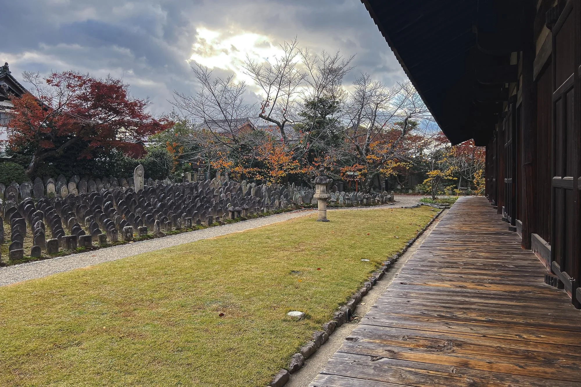 A serene Japanese temple garden with a wooden walkway on the right, manicured grass and trees with autumn-colored leaves, stone lantern and stones on the left, under a cloudy sky.