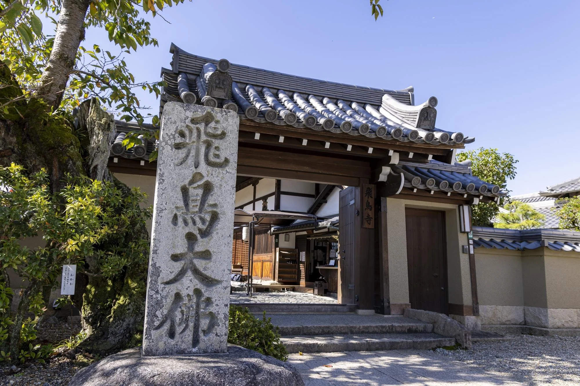 Traditional Japanese temple entrance with a stone pillar inscribed with Japanese characters and a wooden gate, surrounded by trees and greenery.