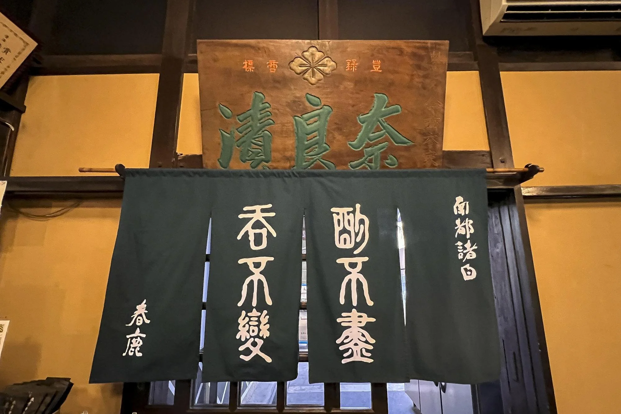 Traditional Japanese entrance curtain with white kanji characters on a dark green background, hanging in front of a doorway, with a wooden sign with green kanji characters above it.