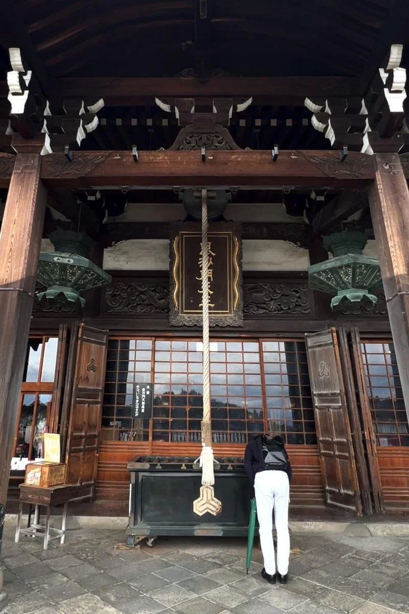 A person with a backpack and white pants bowing at a traditional Japanese shrine with a large hanging rope and bell.