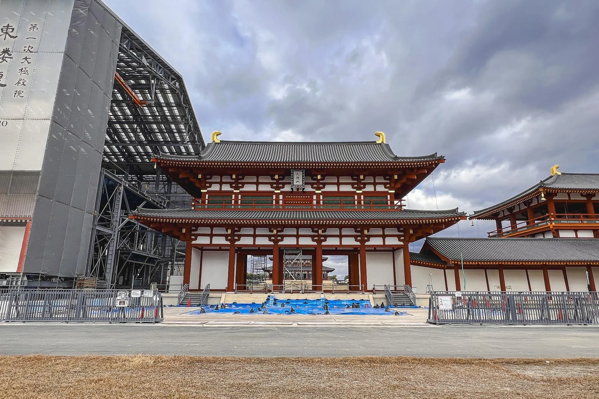 A traditional Japanese temple with red and white walls, ornate roofing, and scaffolding on the left side indicating ongoing construction or renovation. The area in front is fenced off with signs and cones, and the sky above is cloudy.