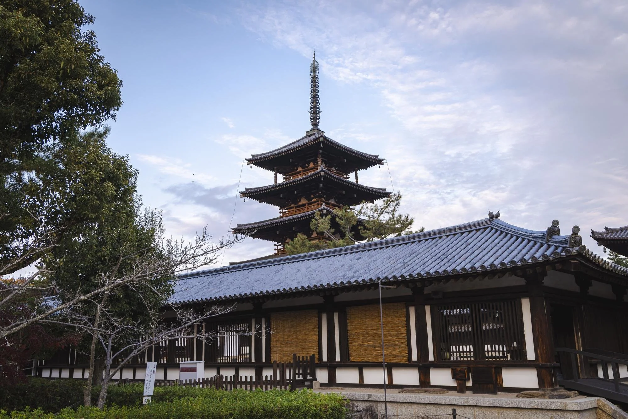 Traditional Japanese temple with a multi-tiered pagoda and tiled roof, surrounded by trees under a partly cloudy sky.