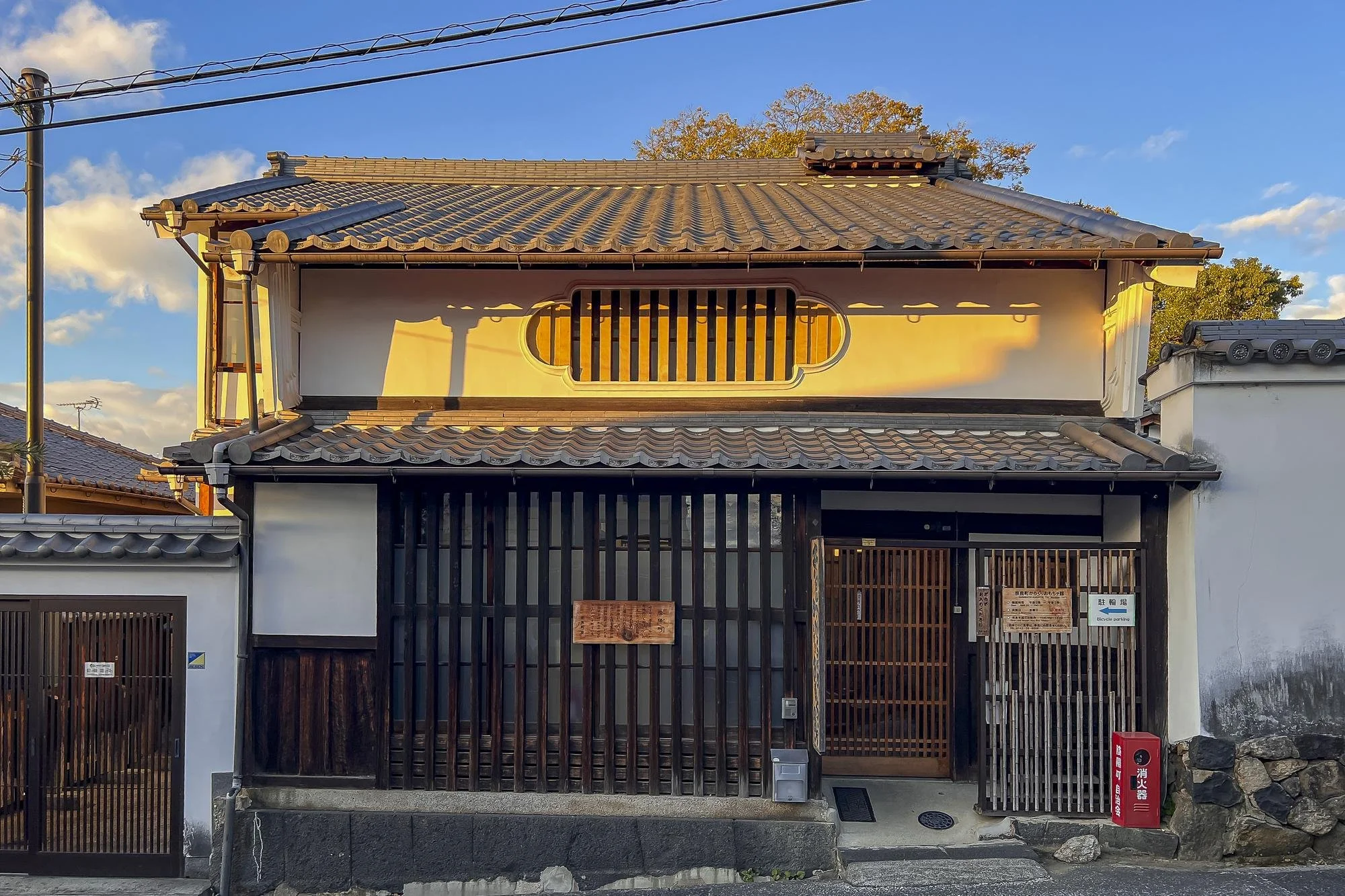 Traditional Japanese house with a tiled roof, wooden sliding gates, and a small stone path at the entrance, illuminated by sunlight at sunset.