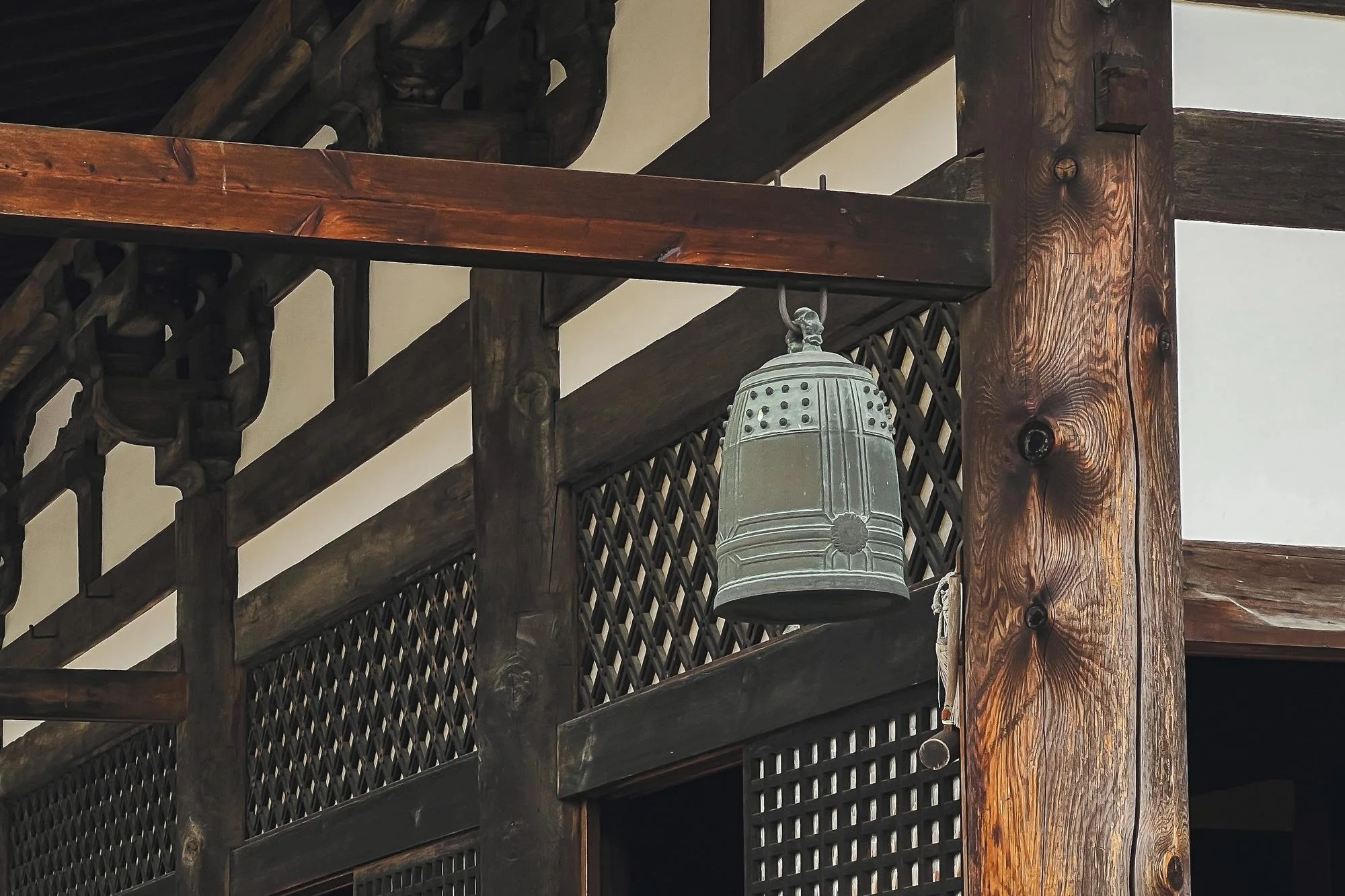 A black metal bell hanging from a wooden structure inside a building with wooden beams and latticework.