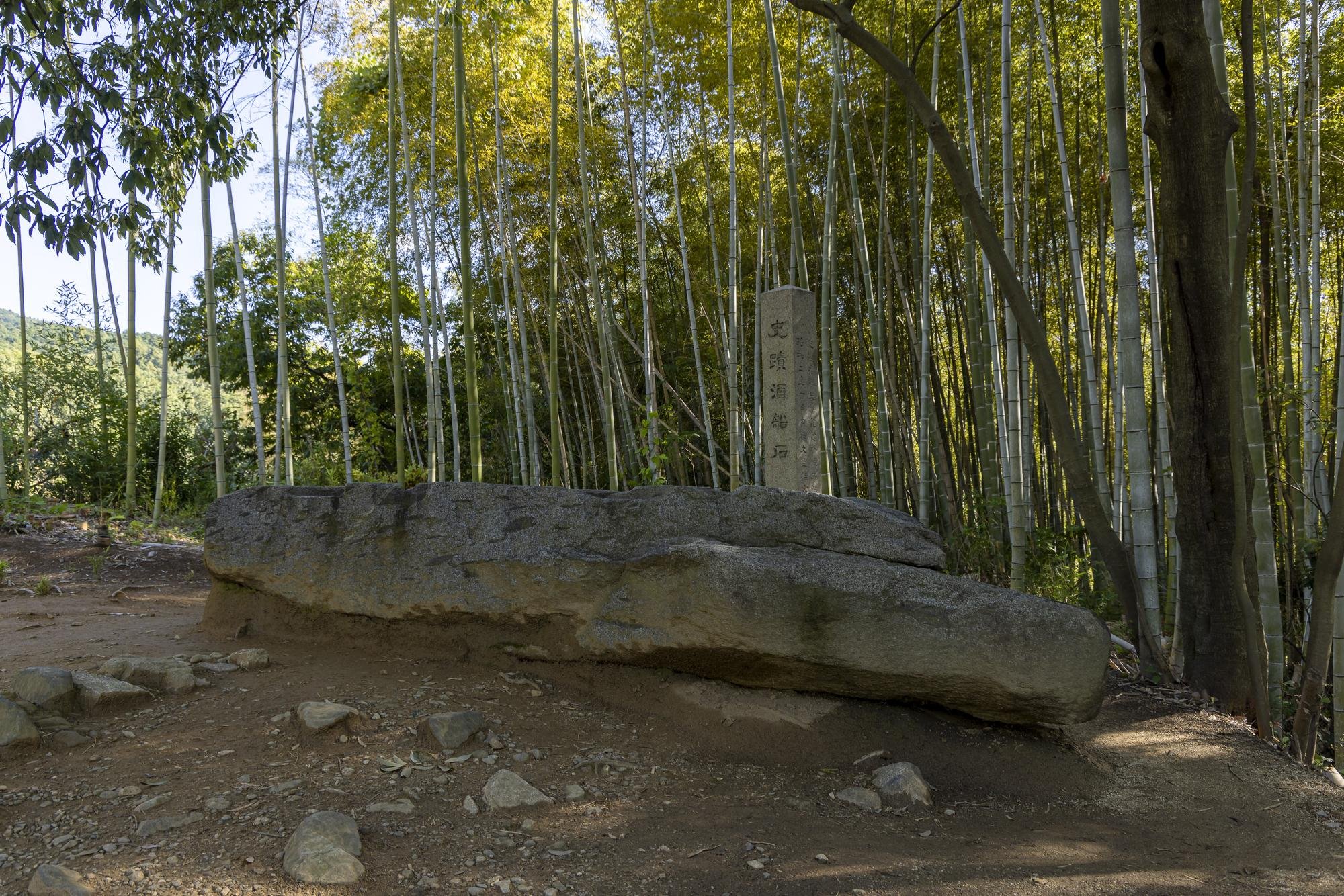 Large flat rock with an upright stone marker behind it, located in a bamboo forest with sunlight filtering through tall bamboo stalks and trees.
