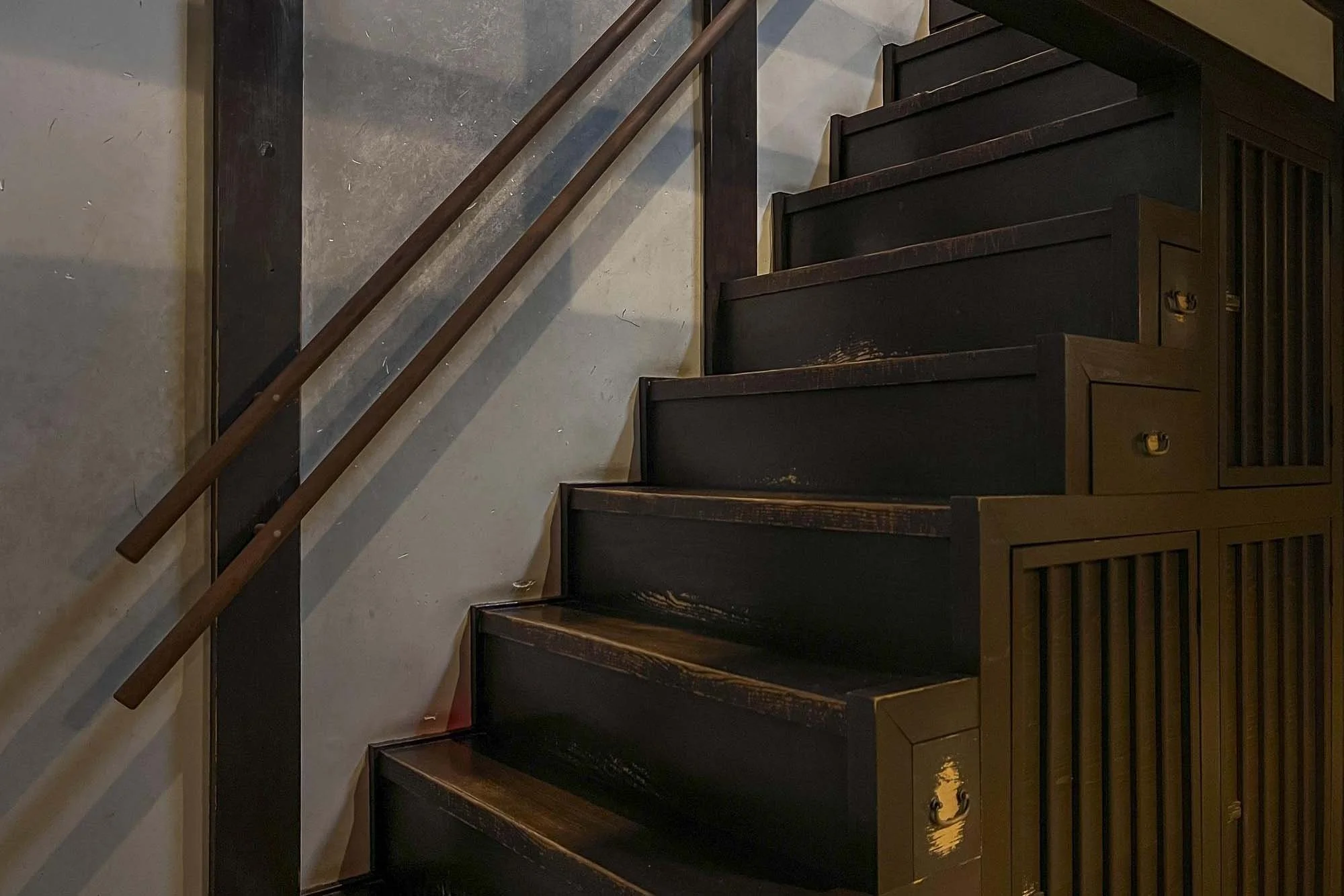 Dark wooden staircase with a handrail along the left side, ascending alongside a light-colored wall.