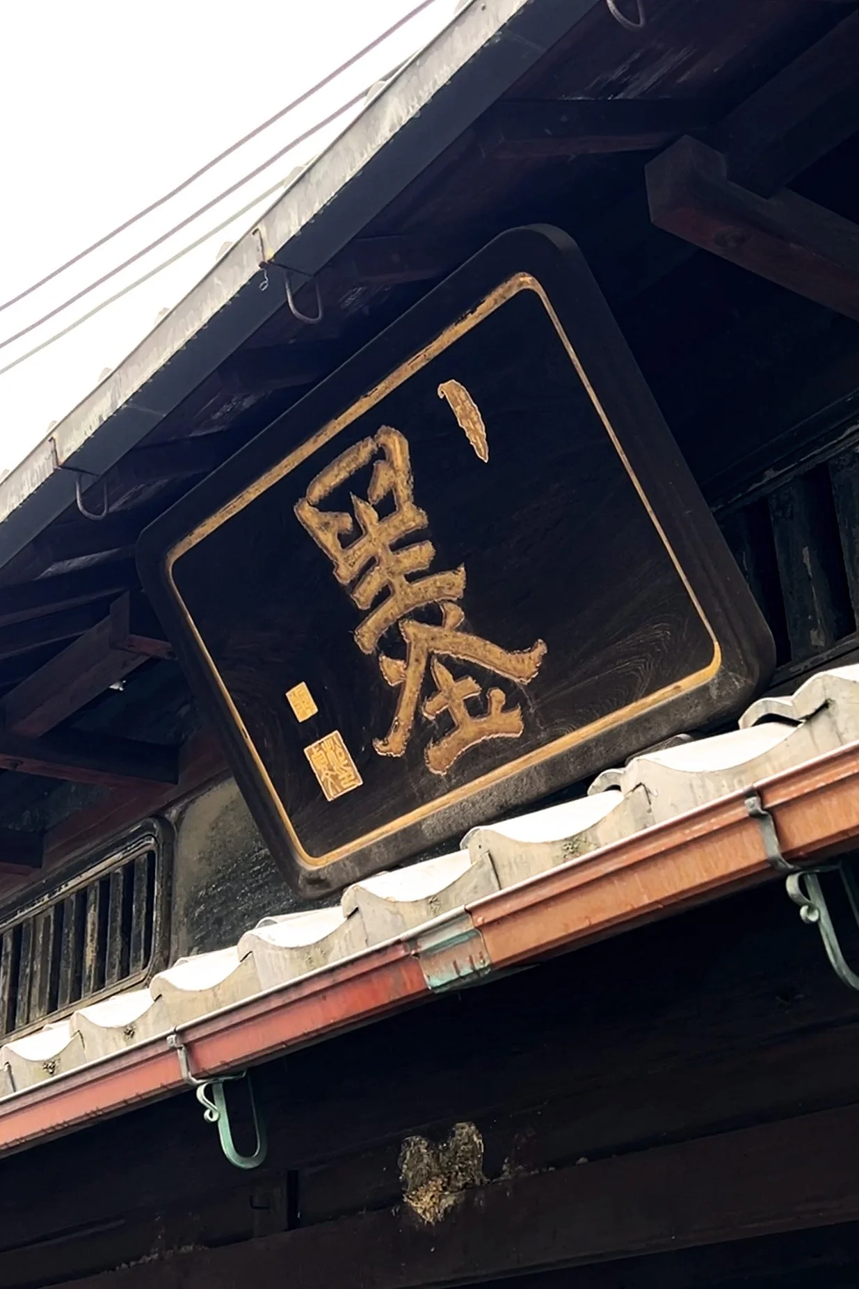 Black square sign with gold Japanese calligraphy characters hanging under a wooden roof, with a tile roof and gutter visible.