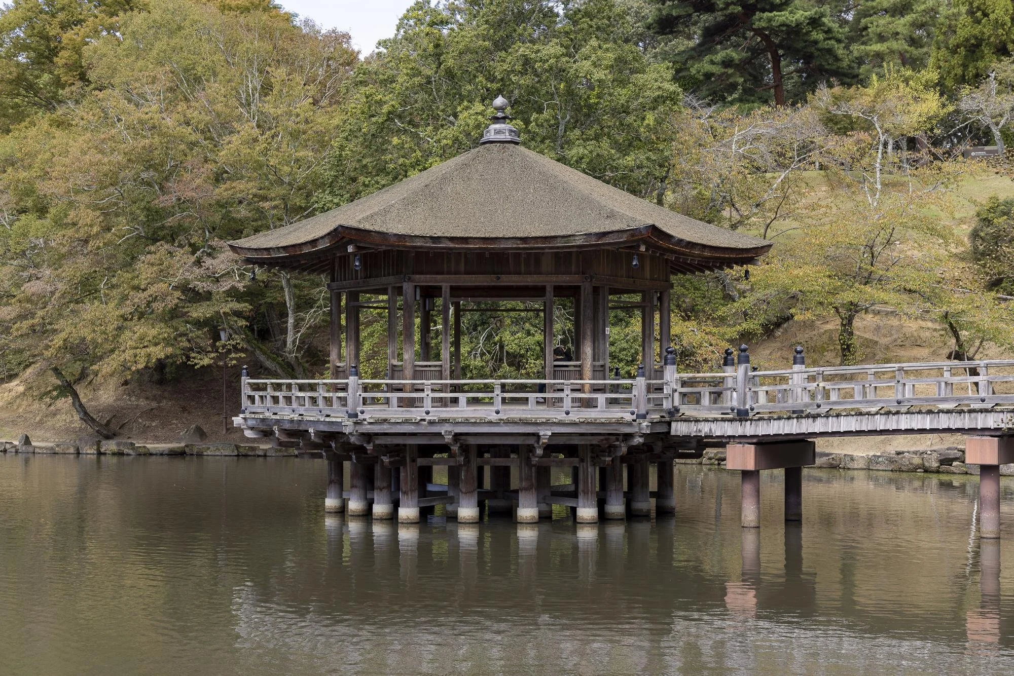 A traditional Japanese-style wooden pavilion with a thatched roof on stilts over a calm body of water, surrounded by trees with autumn foliage.