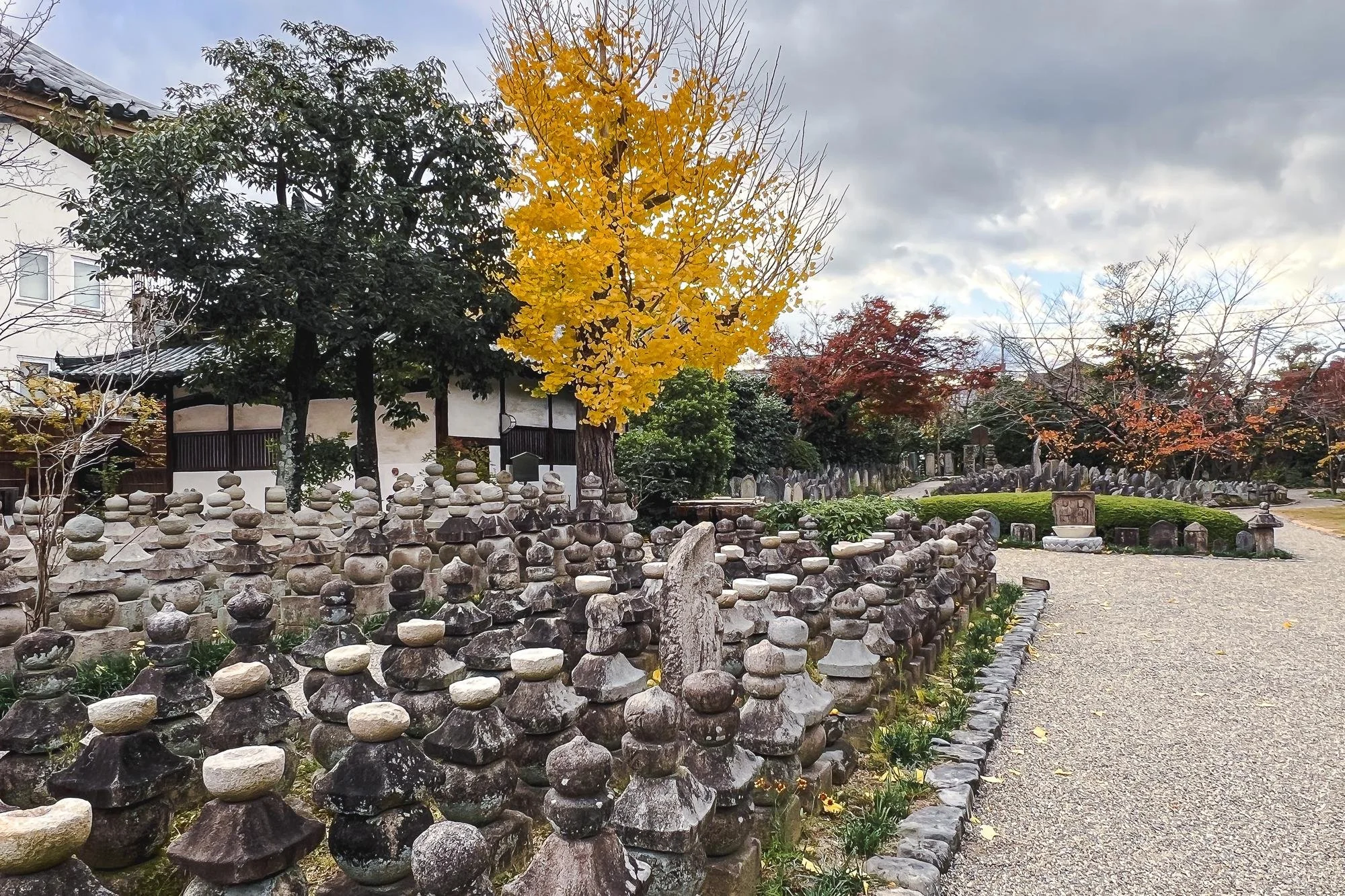 A Japanese garden with numerous small stone pagodas lining a gravel path, trees with autumn foliage in shades of yellow, red, and orange, and a cloudy sky overhead.