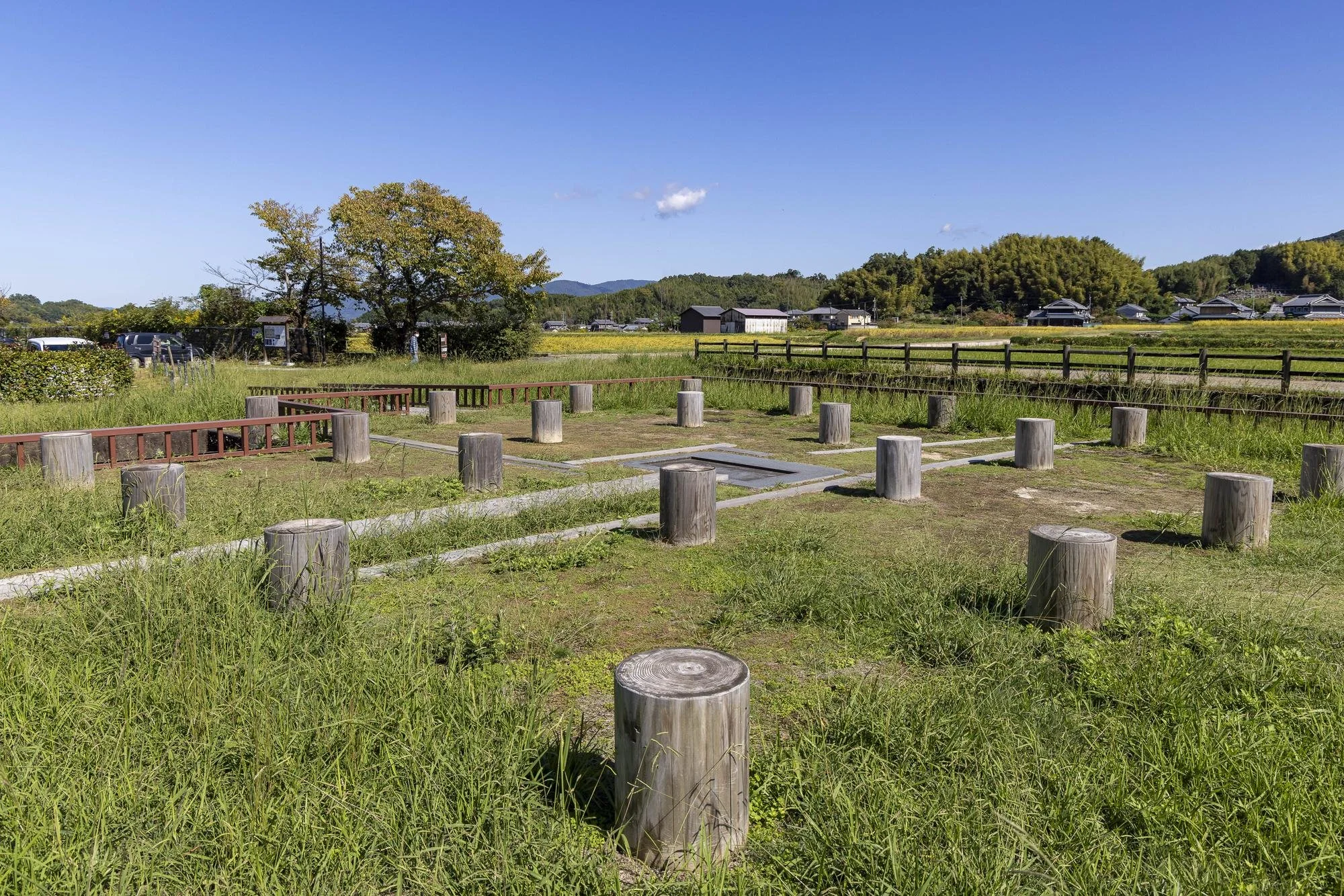 Asuka Water Clock Site: Open grassy area with wooden posts and low fences, trees in the background, clear blue sky, and distant buildings.