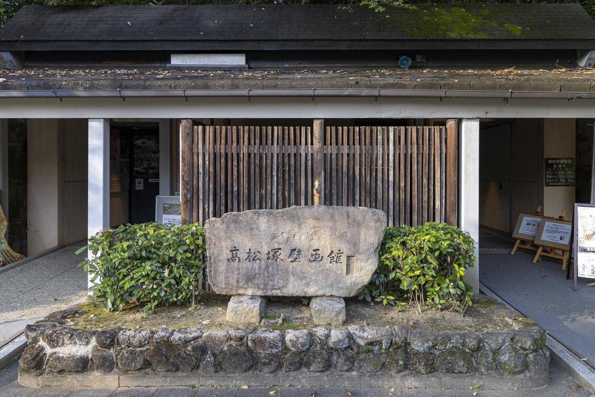 Stone sign with Japanese characters in front of a building entrance, surrounded by small green bushes, with a wooden fence and signs inside the building visible behind.