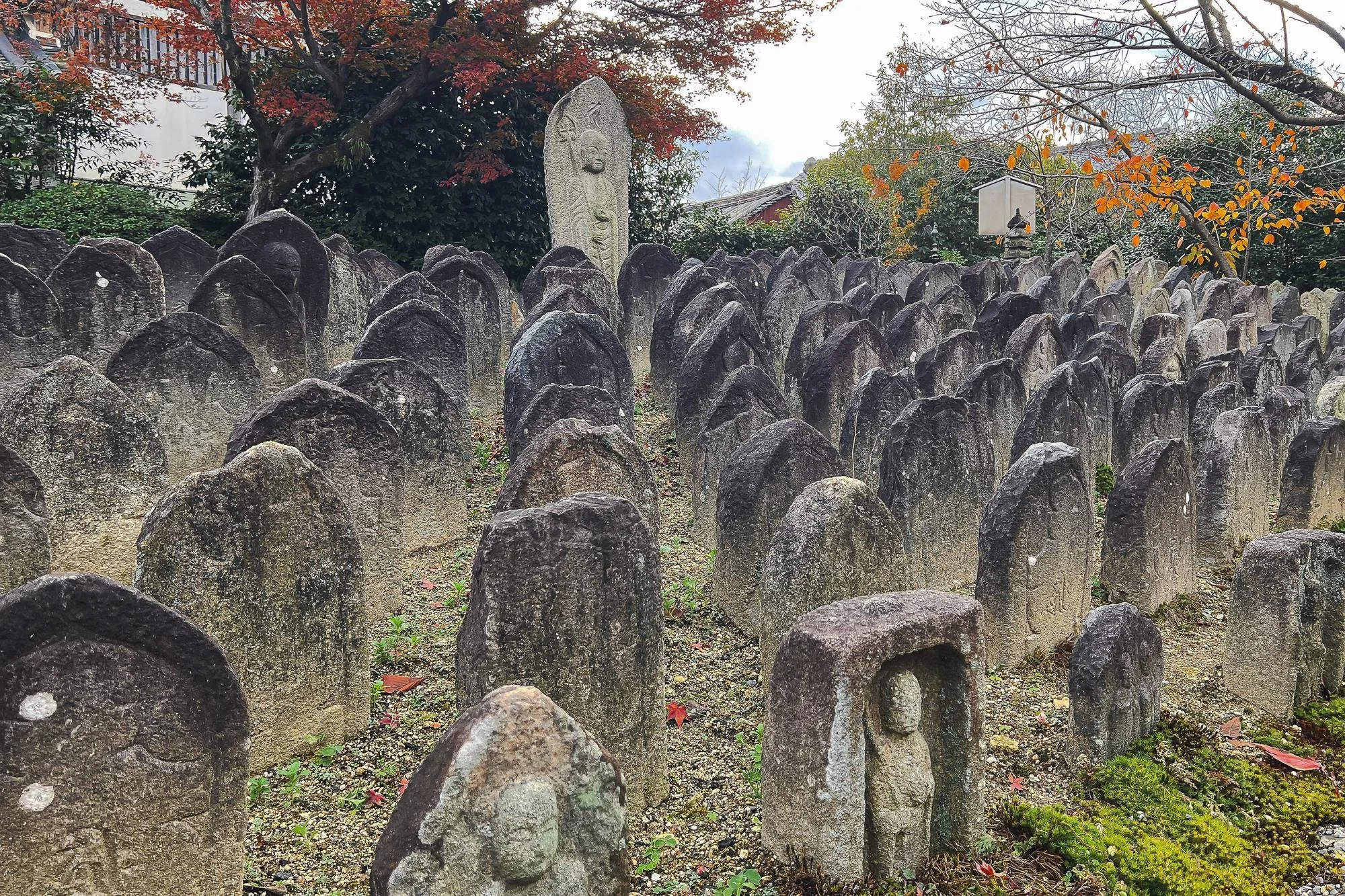 Row of weathered stone grave markers in a cemetery, with some markers featuring carved faces or symbols. In the background, a large carved stone monument, trees with fall foliage, a small lamp post, and a building are visible.
