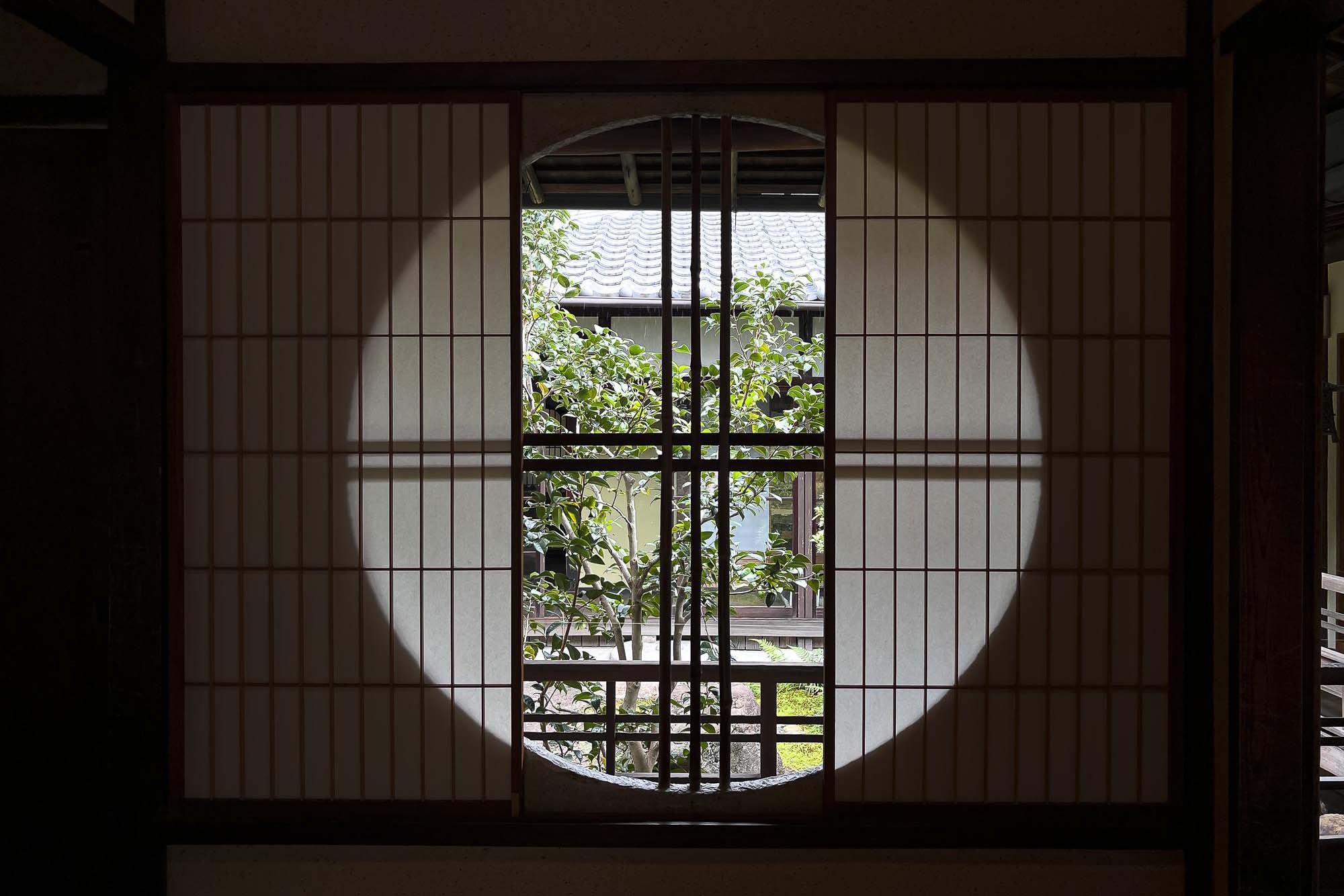 Traditional Japanese shoji sliding paper door with a circular opening, revealing a garden with a tree and a wooden building outside.