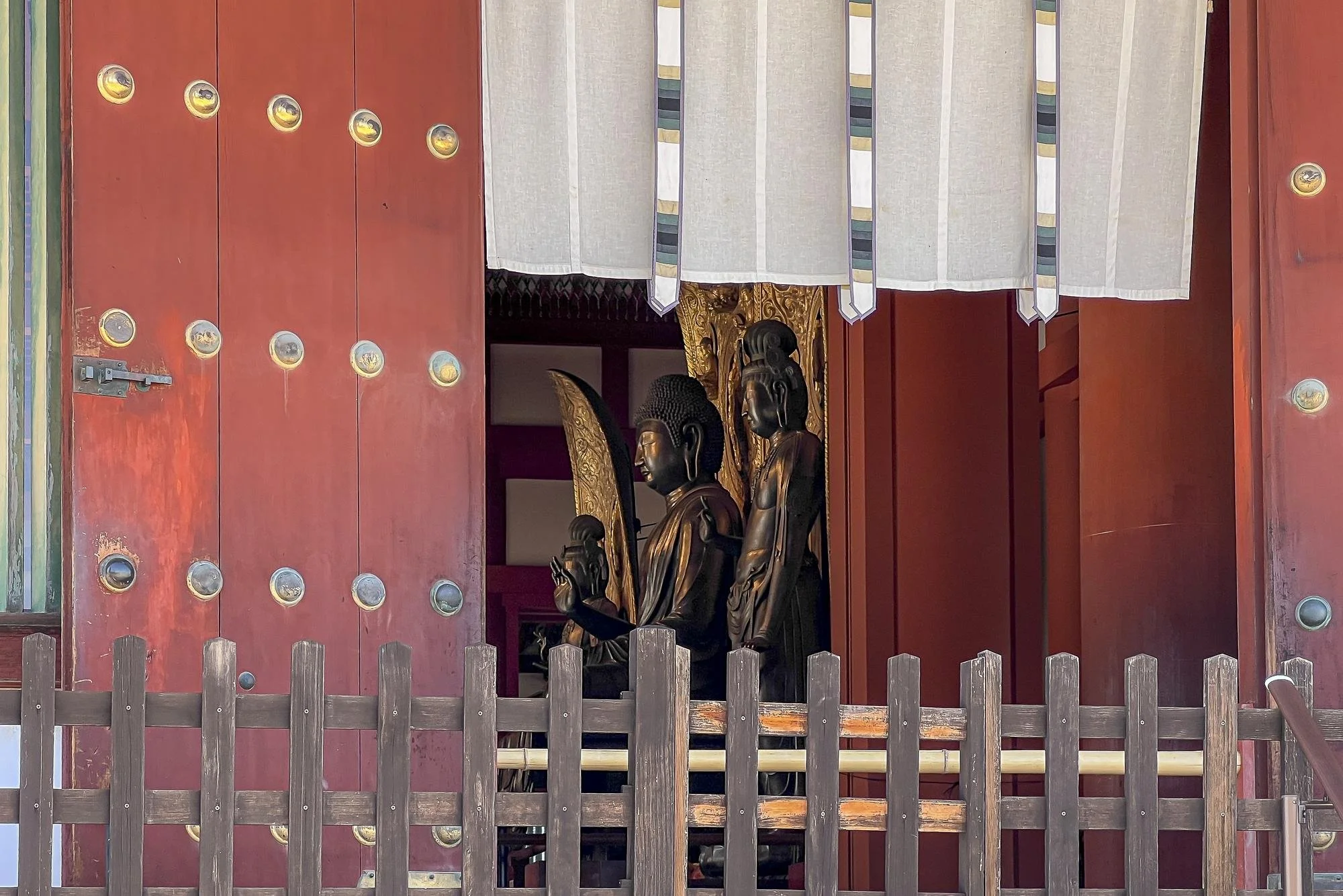 View of traditional Japanese temple with carved wooden statues of Buddha and attendants inside, partially visible through an open red wooden door with circular decorations, surrounded by a railing.