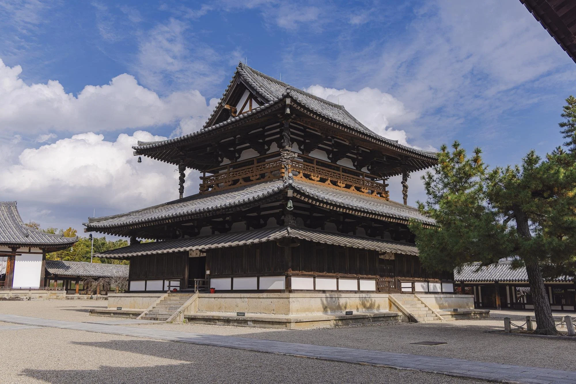 Traditional Japanese temple with multiple tiers, wooden architecture, curved tiled roofs, and surrounding trees under a blue sky with clouds.