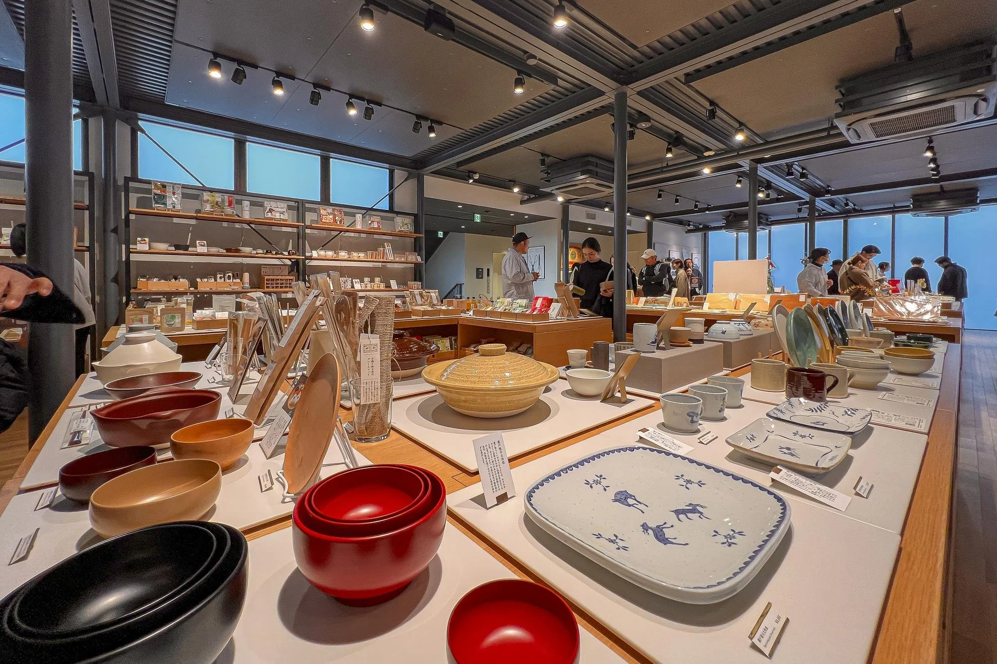 Interior of a store displaying various bowls, plates, and ceramics on white tables, with shoppers browsing and examining items. Large windows in the background let in natural light.