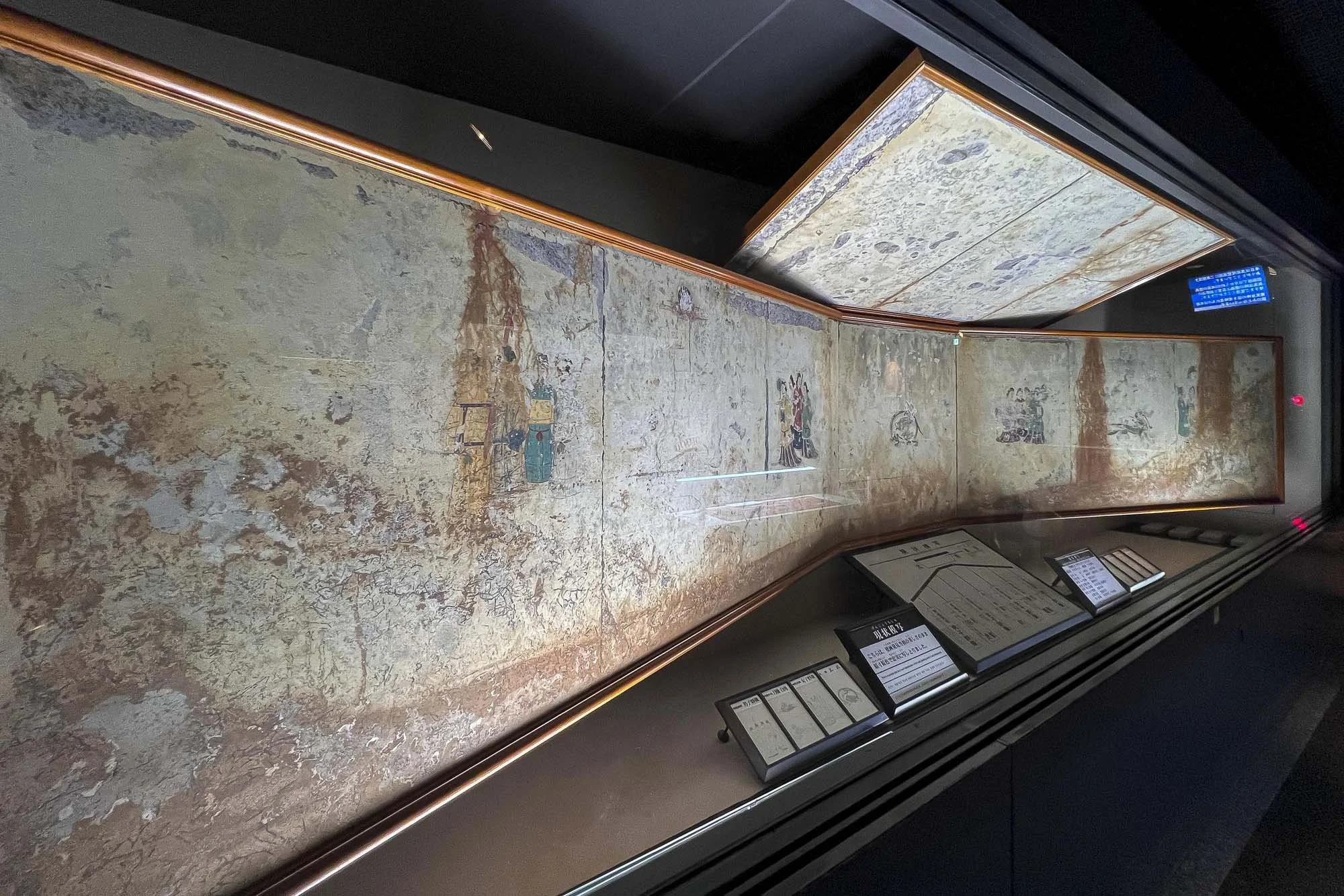 Ancient mural painting displayed in a glass case at a museum, with informational plaques below.
