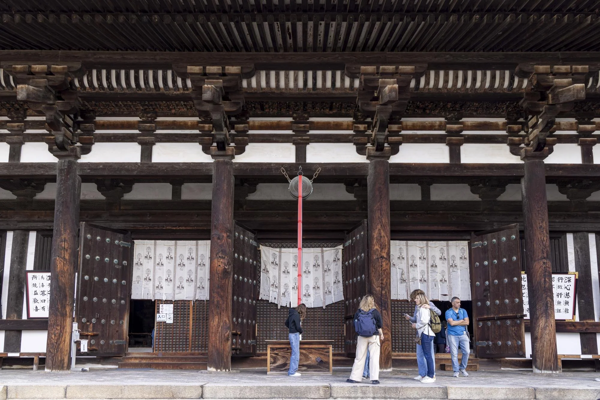 Five people standing in front of a traditional wooden Japanese building with large doors and hanging curtains, some looking at their phones or talking.