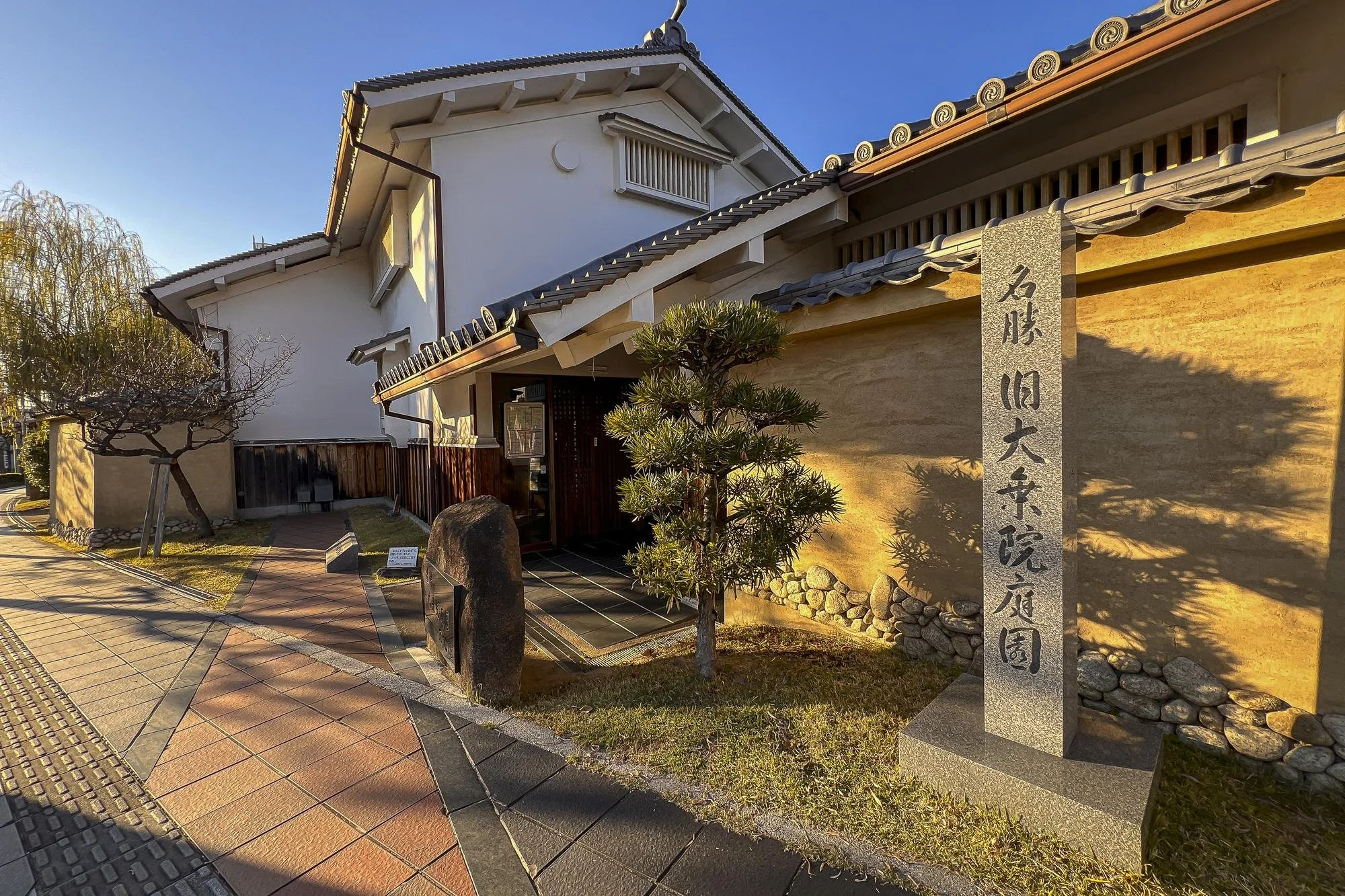 Traditional Japanese building with a tiled roof, a stone sign with Japanese characters, a small tree, and a pathway in sunlight.