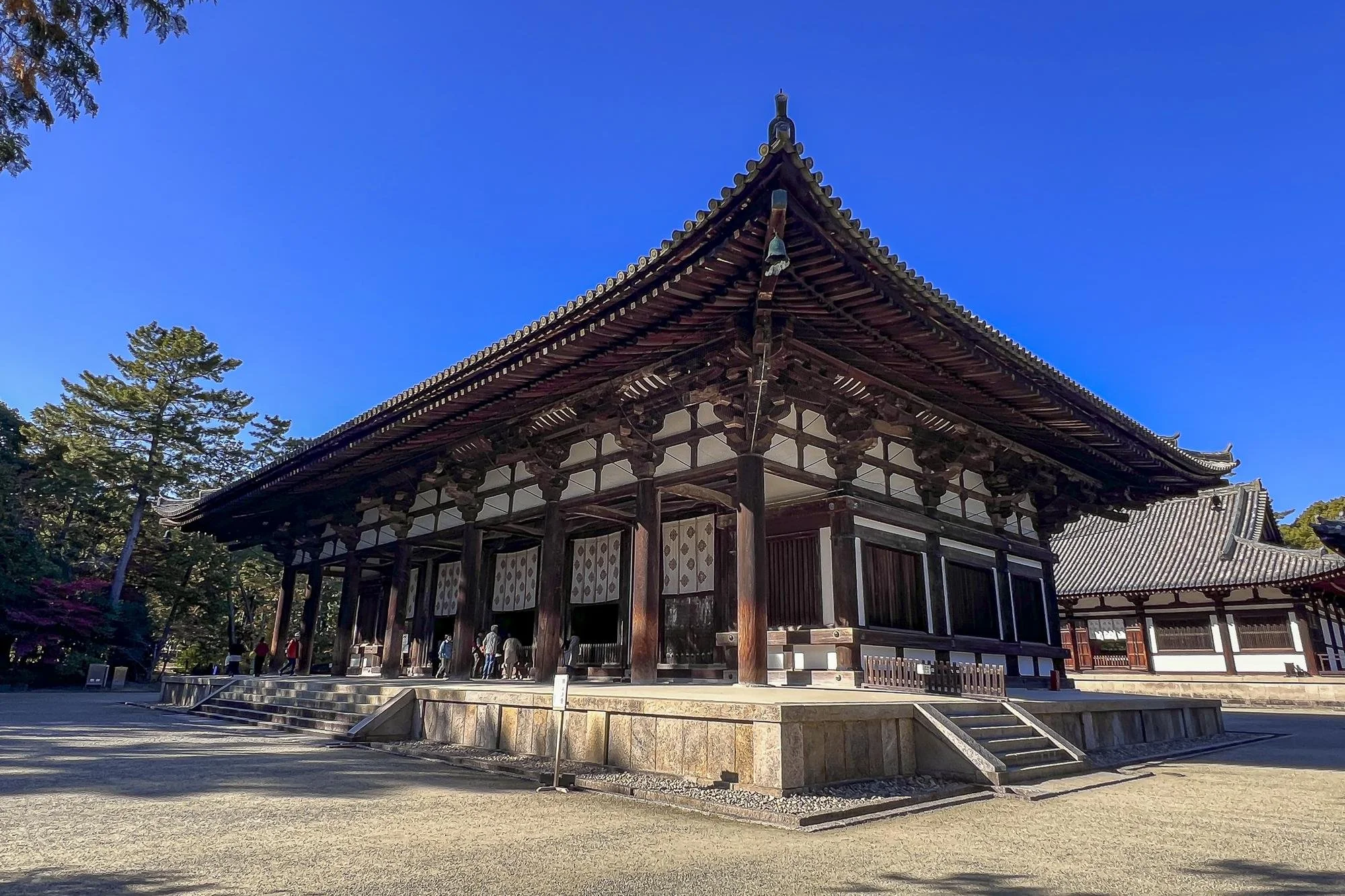 Traditional Japanese temple with wooden structure, tiled roof, and stone steps, surrounded by trees under a clear blue sky.