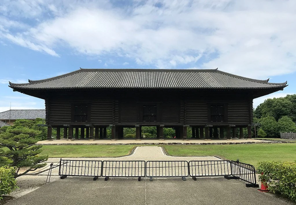 Traditional Japanese wooden building on stilts with a tiled, curved roof, surrounded by greenery and a clear sky.