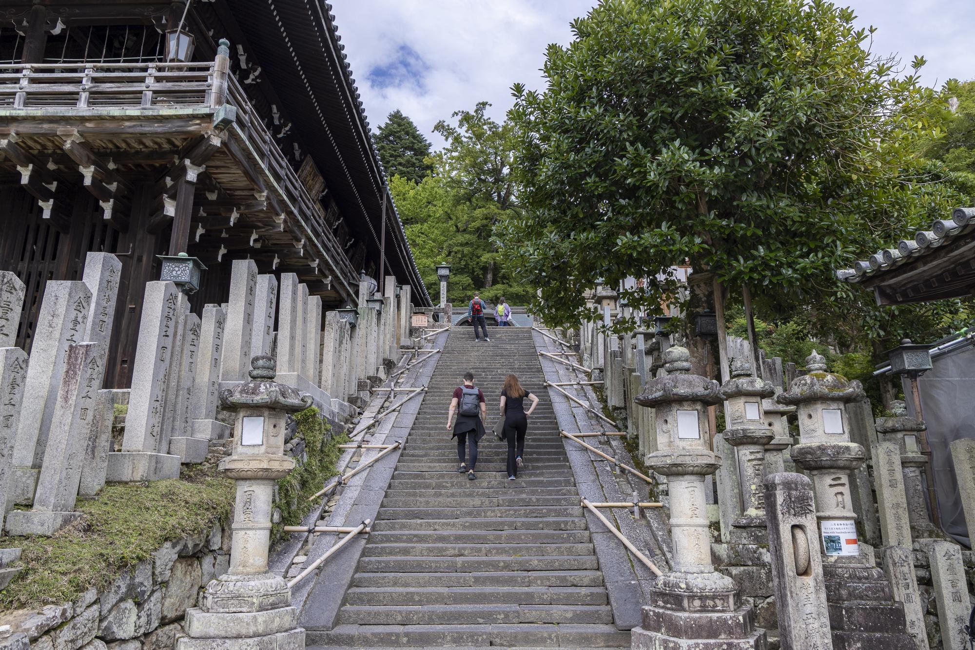 People ascending stone steps leading to a traditional Japanese shrine, flanked by stone lanterns and surrounded by trees.