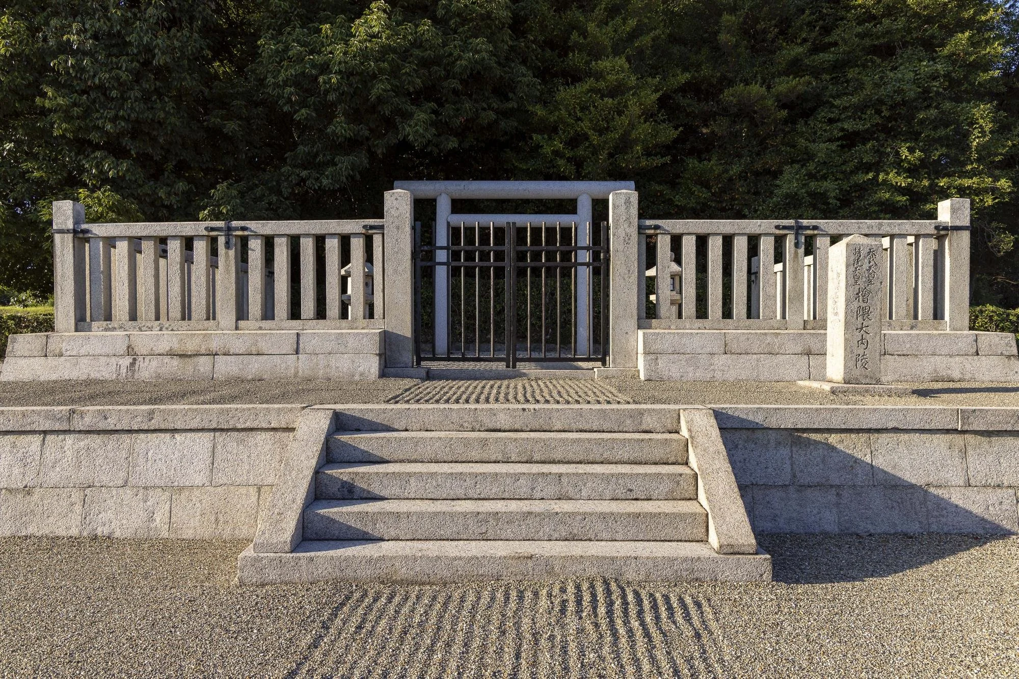 Stone steps leading up to a gate with a wooden fence and a stone pillar with Japanese inscriptions, with trees in the background.