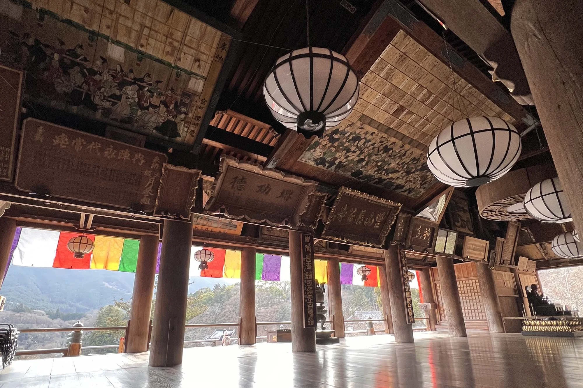 Hase-dera Temple: Interior of a traditional wooden structure with hanging paper lanterns, colorful banners, and hanging scrolls or artwork, overlooking a scenic mountain view outside.