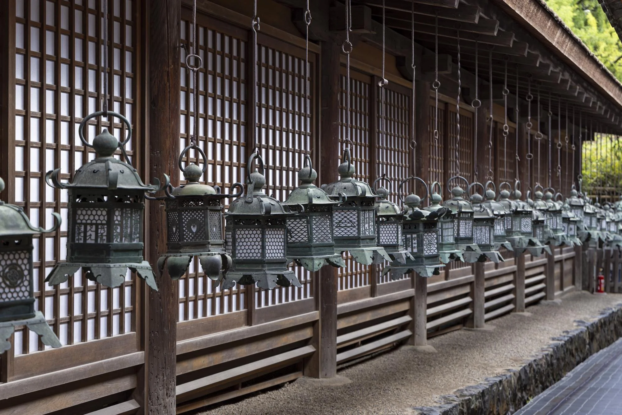 Several traditional metal lanterns hanging in a row in front of a wooden lattice wall at a Japanese temple or shrine.