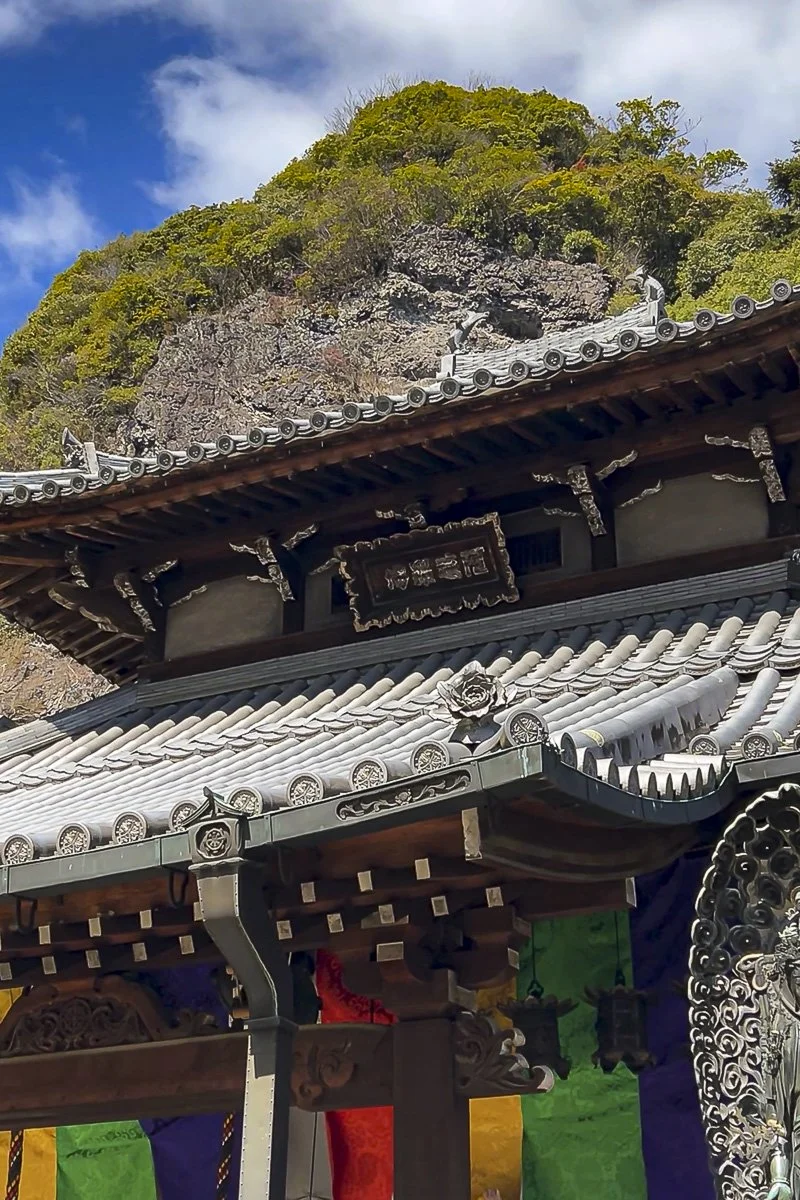 Roof of a traditional Japanese temple with ornate tiles and decorations, set against a rocky hill with green foliage and a partly cloudy sky in the background.