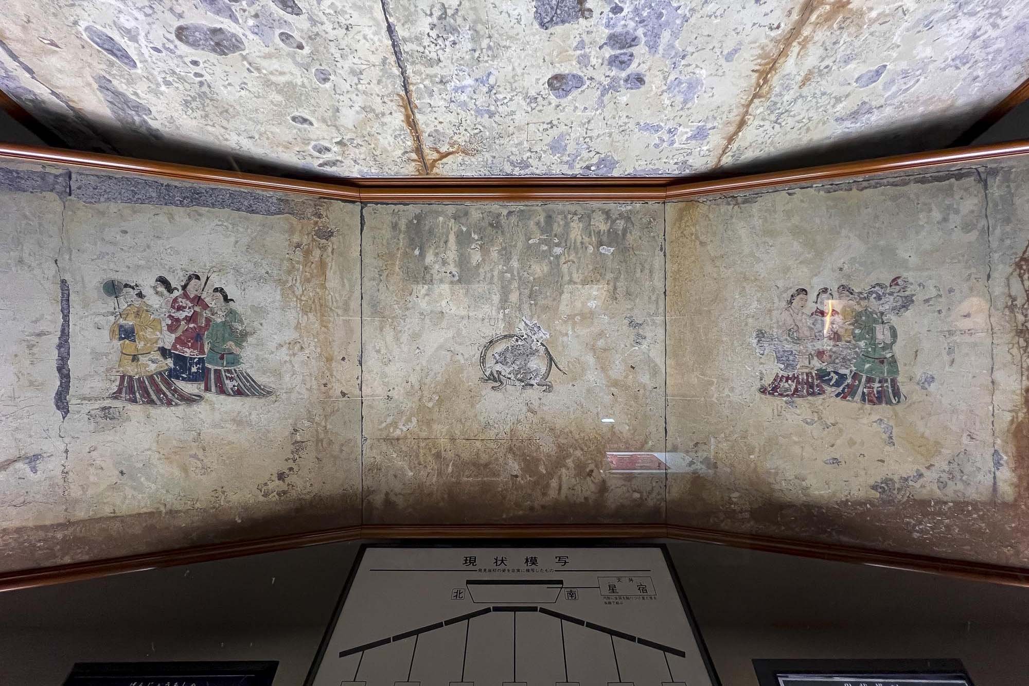 Ancient mural painting depicting three groups of people in traditional attire, surrounded by a muted, weathered background. The mural is behind glass in a museum setting.