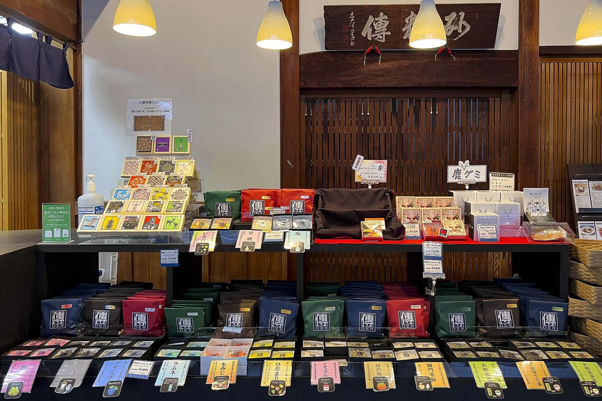 Display of packaged teas and snacks in a shop with Japanese signs, including colorful tea bags and boxes, and a bottle of hand sanitizer.