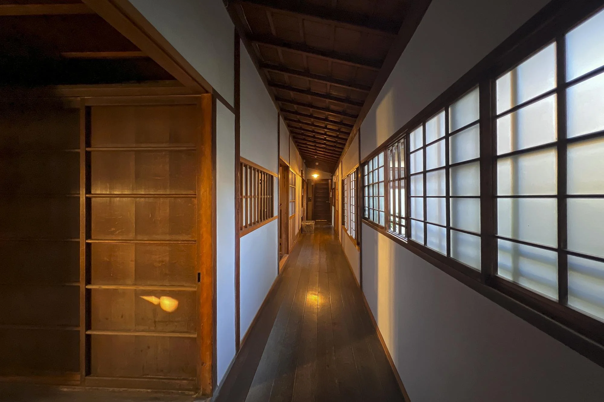 A long wooden hallway with traditional Japanese sliding paper doors and windows, warm lighting, and a polished wooden floor.