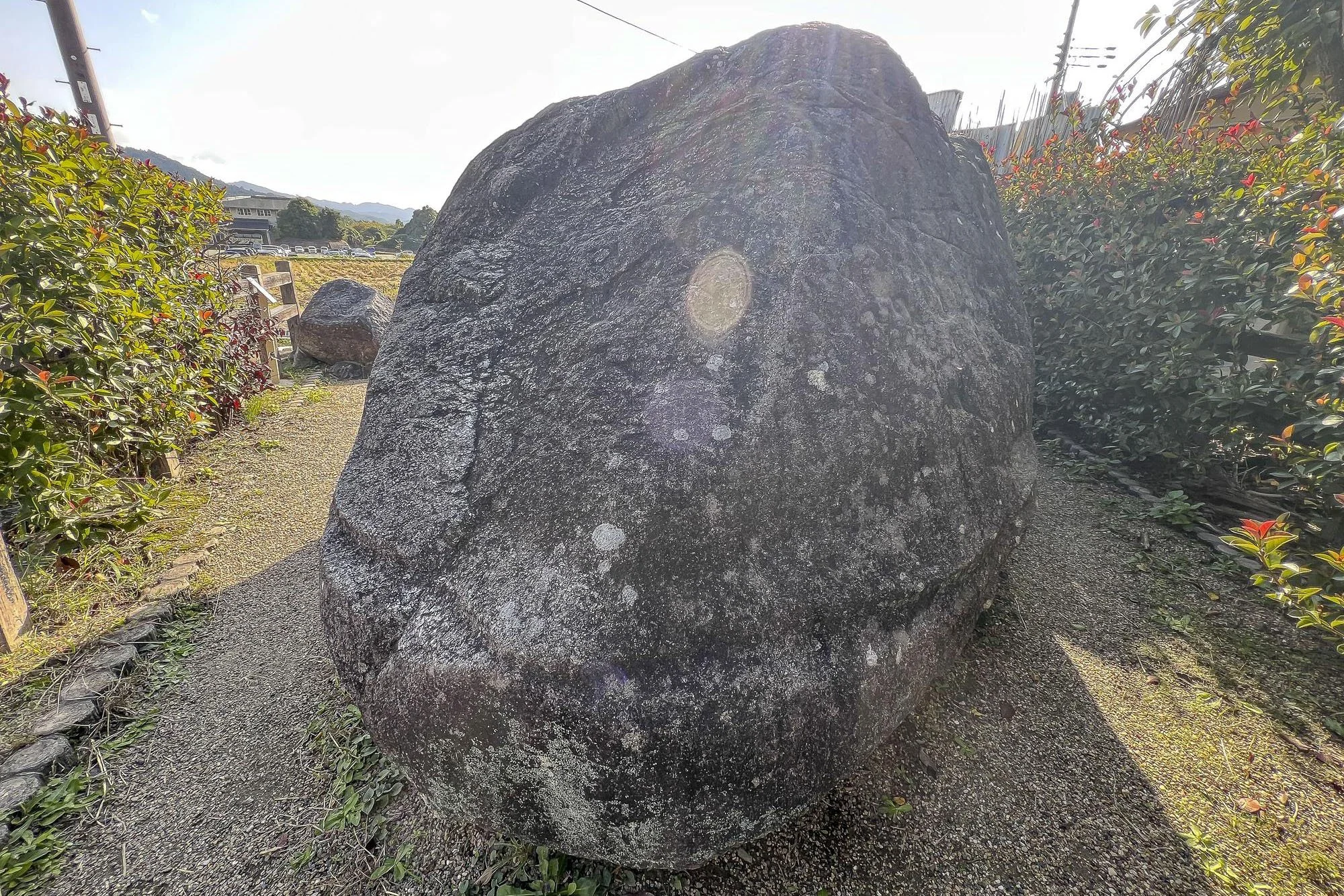 A large, rounded boulder surrounded by greenery on both sides, with a dirt path in front, under bright sunlight.