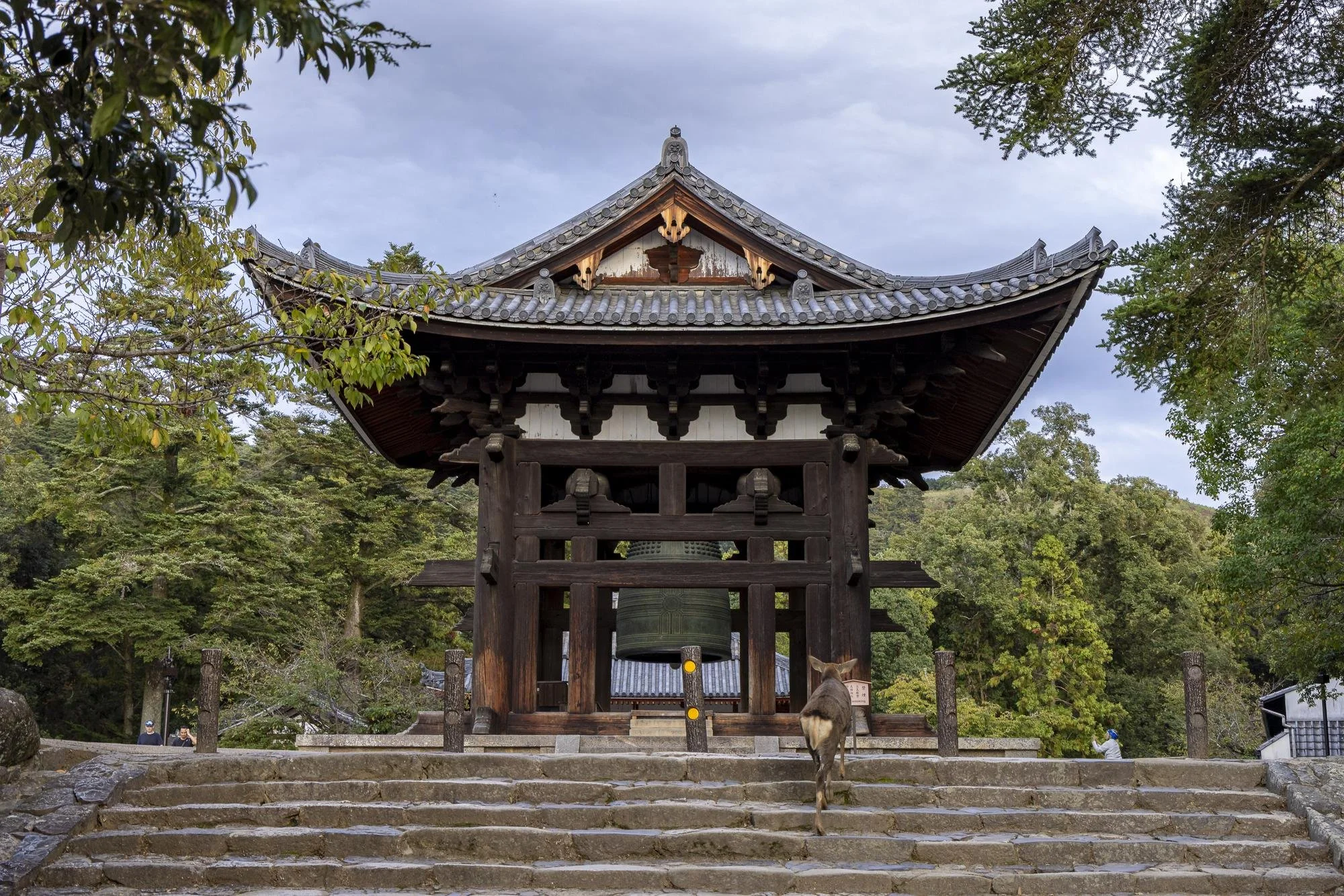 A traditional wooden temple with a large bell in the center, situated on stone steps, surrounded by green trees and a cloudy sky.