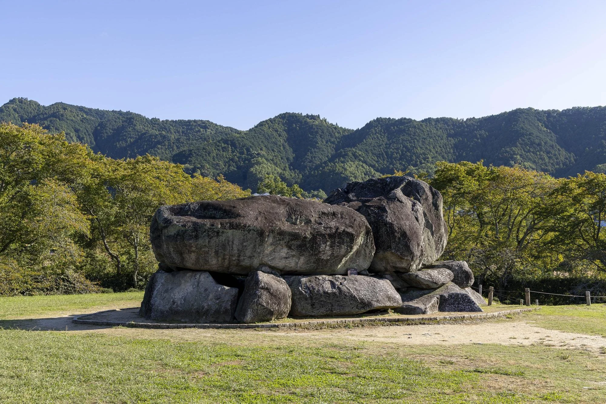 Large rocks stacked on a grassy area with trees and green mountain range in the background under a clear blue sky.
