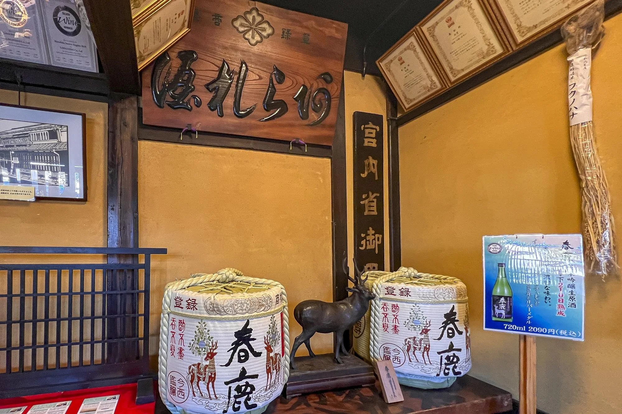 Decorative display with two large sake barrels, a bronze reindeer statue, and various Japanese signs and certificates on the wall inside a restaurant or store.