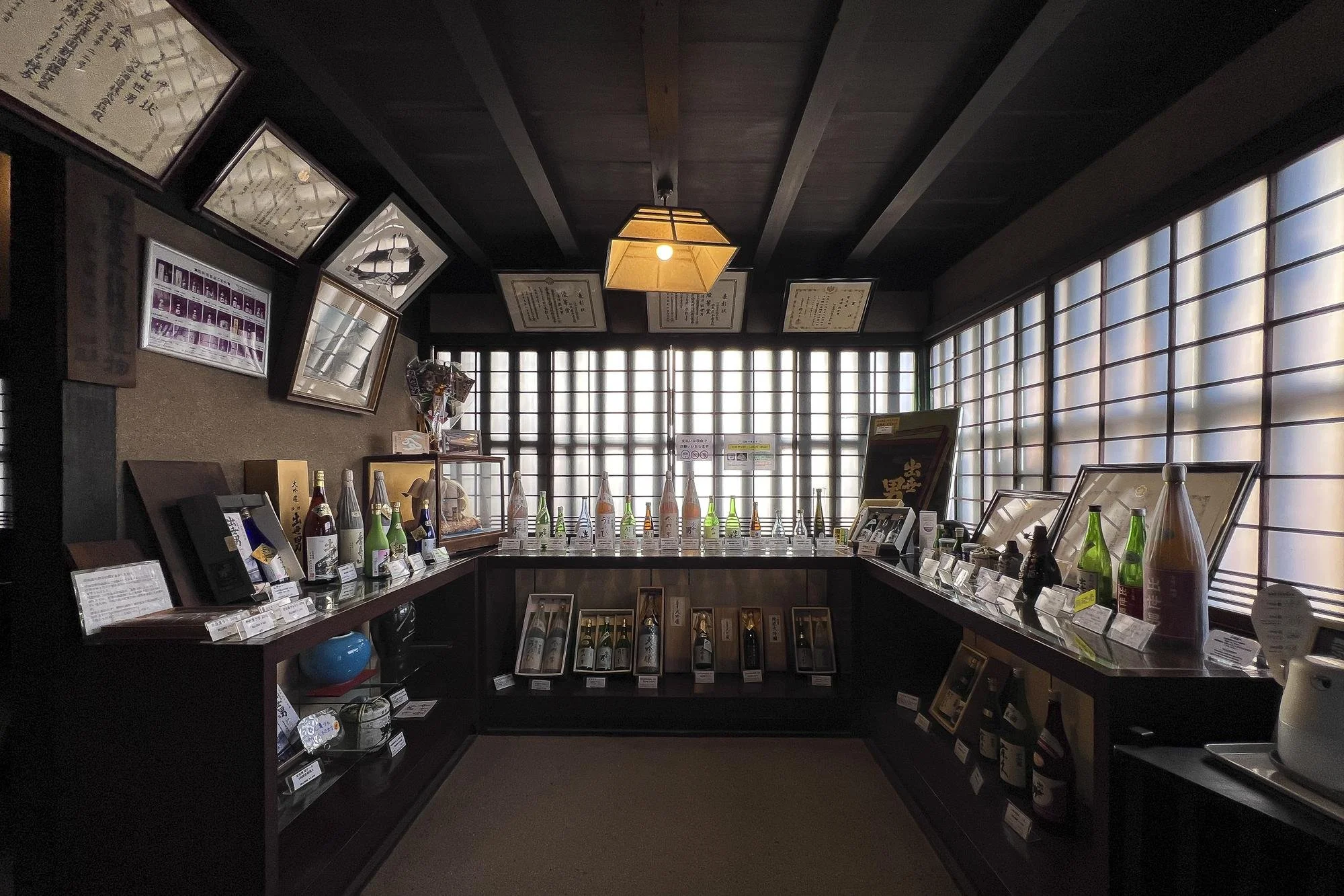 An interior of a Japanese sake shop with bottles of sake displayed on shelves, framed pictures, and certificates on the walls, illuminated by traditional shoji screens and a hanging paper lantern.