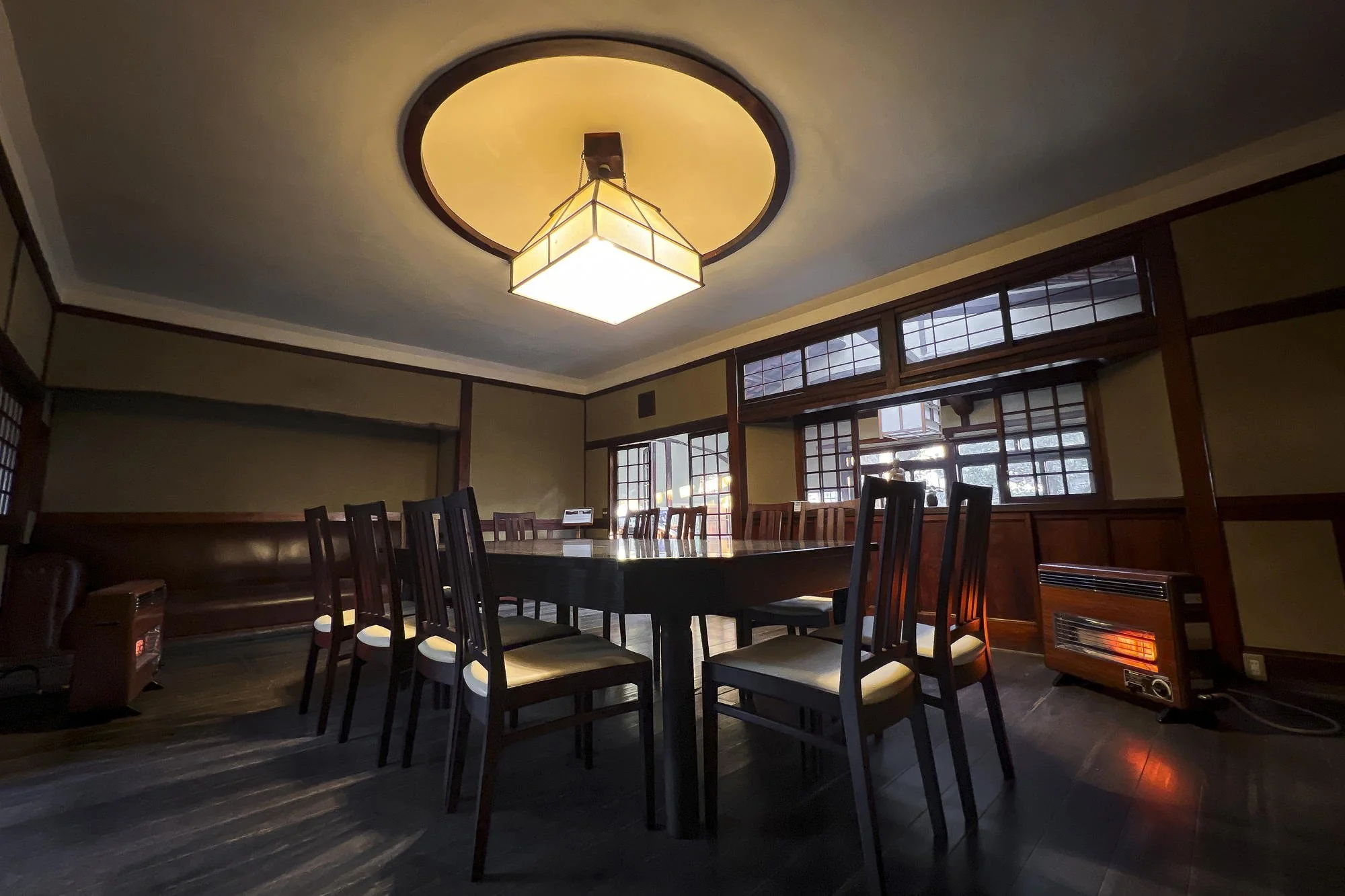 Empty room with a large dining table, wooden chairs, and traditional Japanese windows, illuminated by a ceiling light and a space heater.