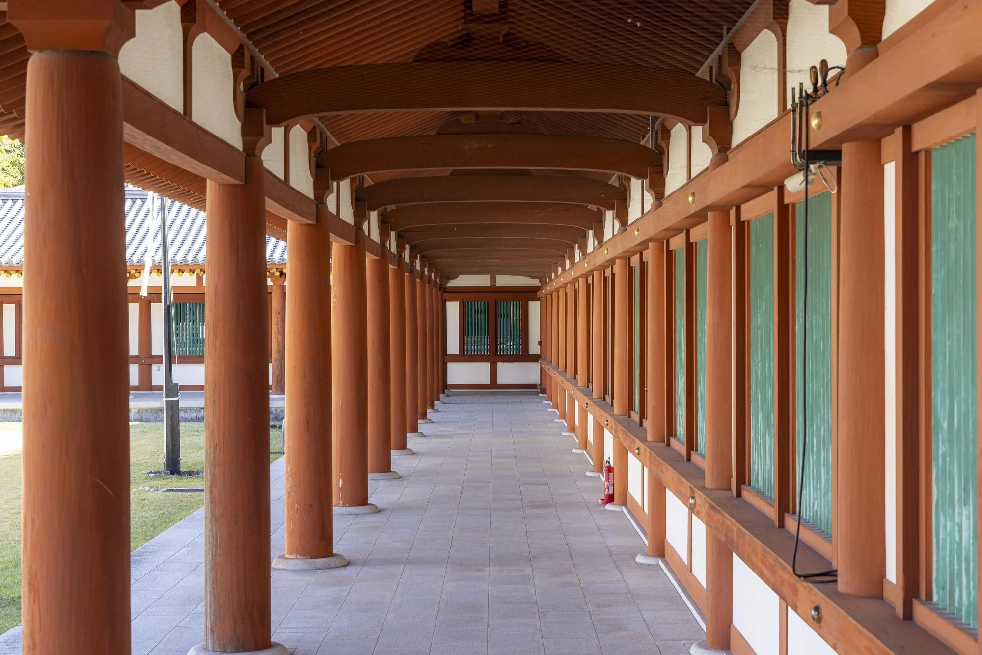A corridor with wooden pillars and a curved wooden roof, leading to a door with green window panels in a traditional East Asian building.