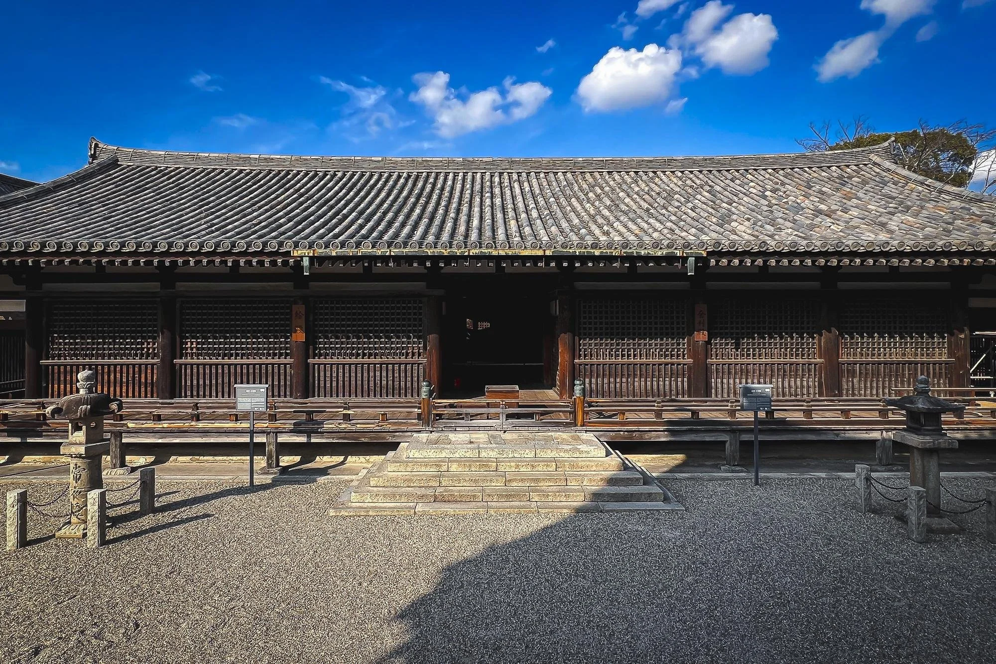 Traditional Japanese wooden building with a tiled roof, steps leading to the entrance, and a gravel courtyard under a blue sky with clouds.