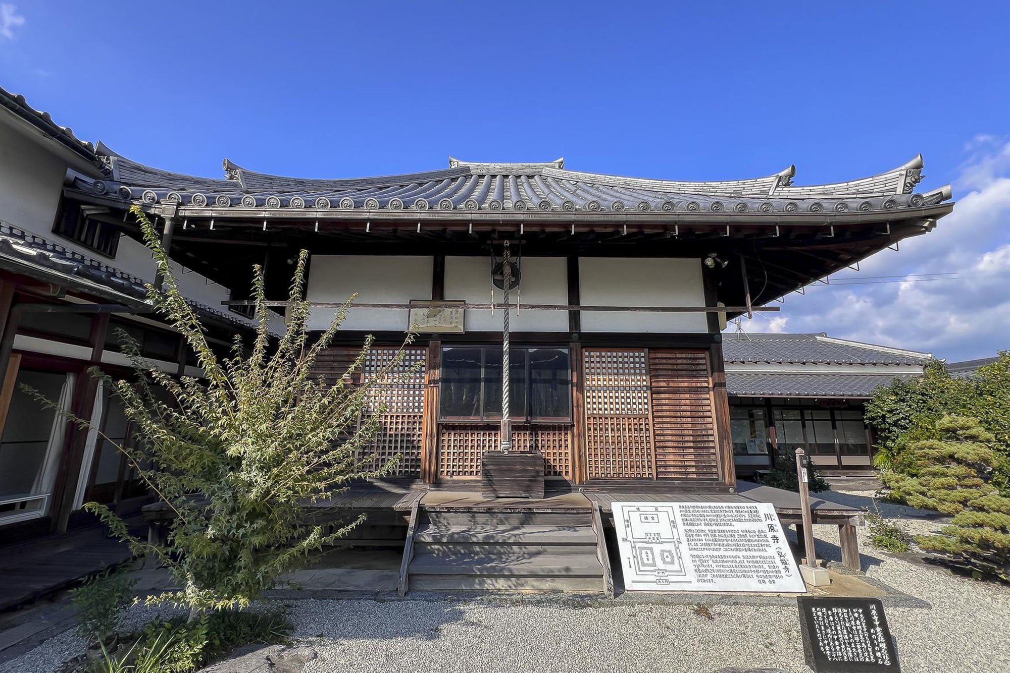 Kawahara-dera Temple: Traditional Japanese building with a wooden exterior, tiled roof, and a shimenawa rope hanging in the center, set against a blue sky, with informational signs in front.